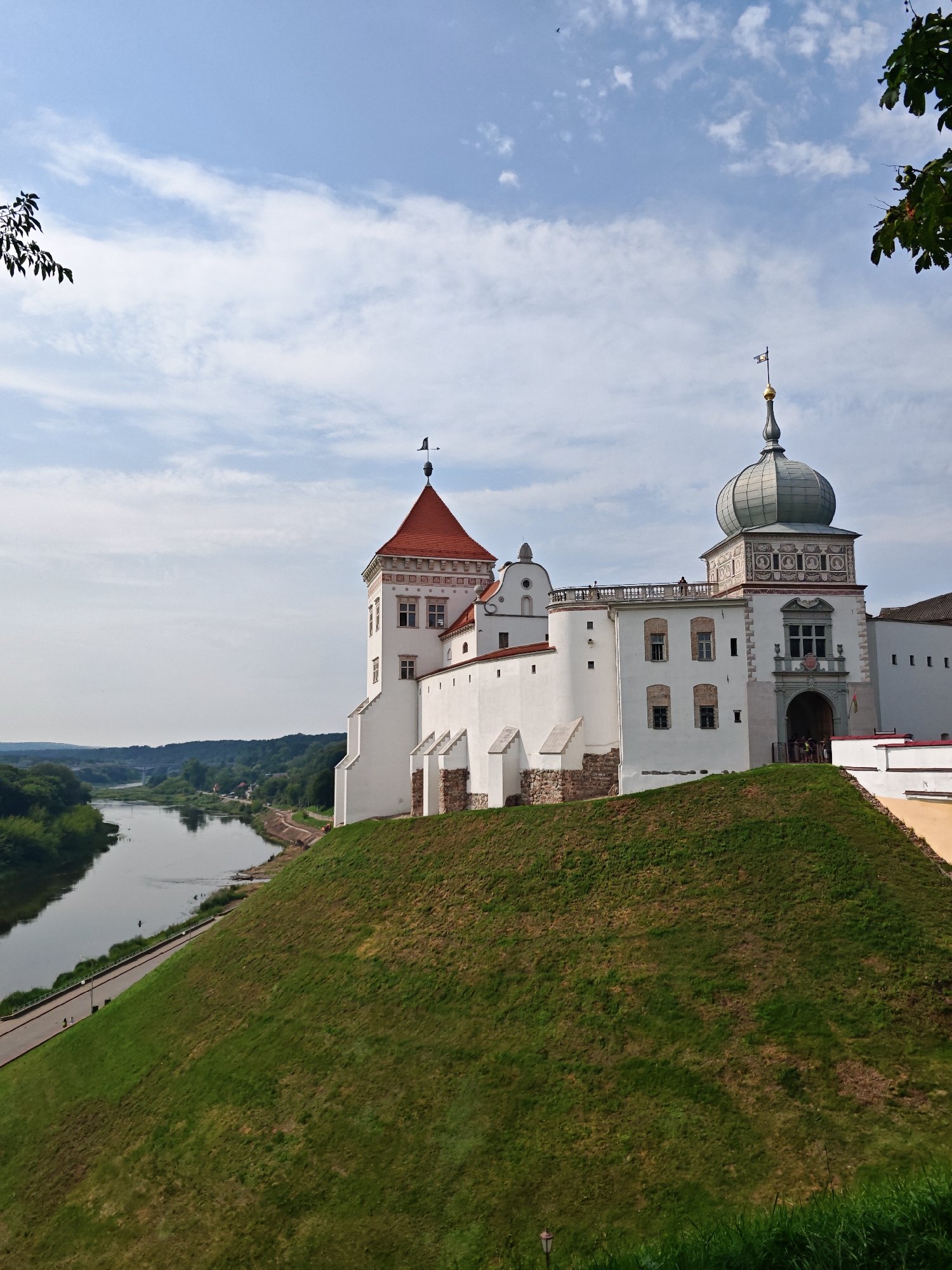 This is a photograph of the Old Castle in Hrodna, Belarus, taken on a sunny day with a partly cloudy sky. The castle is a large, white stone structure with red-tiled roofs and a distinctive green dome. It sits atop a grassy hill, overlooking a wide river.
The castle complex features multiple towers and buildings, with architectural details that suggest a long history. A prominent tower on the right has a decorative façade with roundels and a golden spire topped with a weather vane. Another tower to the left has a conical red roof and a weather vane. The castle walls are accented with stone supports.
Below the castle, a paved path runs along the riverbank, bordered by lush green trees and foliage. The river itself is wide and calm, reflecting the sky. In the distance, a bridge spans the river, and more trees and buildings are visible on the far bank, suggesting a town or city.
There are a few people visible in the scene. Some are walking on the path by the river, while a small group is gathered near the entrance to the castle. A single person can be seen on a balcony on the right side of the castle. A lone figure is also present on the path near the river. A small black bird or insect is visible flying in the sky.