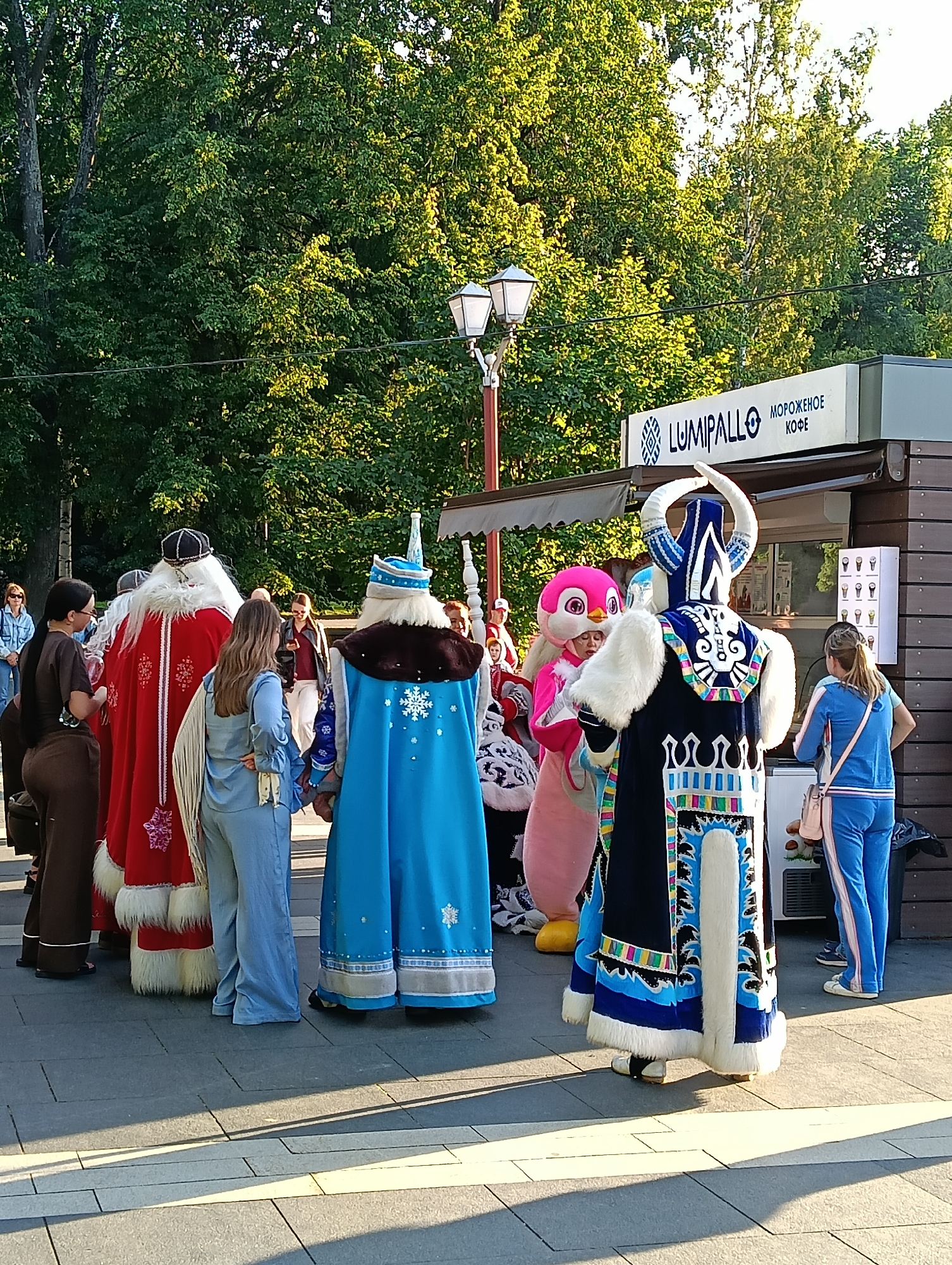 In Petrozavodsk, Russia, on a paved outdoor area lined with trees, a group of individuals are gathered. The scene captures a summer day with dappled sunlight casting shadows. In the foreground, people are dressed in costumes. One person wears a red cloak with white trim and fur accents, resembling Ded Moroz, a Russian winter folklore character. Another person is clad in a blue costume with white fur trim and a snowflake emblem, possibly Snegurochka, Ded Moroz's granddaughter. A pink bird-like mascot costume is also visible, as is a person in a dark blue, ornate costume with white fur and a helmet adorned with horns.
Several other people are present, some in everyday clothing. A woman in a brown outfit stands to the left, observing the costumed figures. Another woman in a blue tracksuit is seen to the right. Behind the costumed individuals, a food stand is present, displaying the logo "LUMIPALLO" and the words "МОРОЖЕНОЕ" (ice cream) and "КОФЕ" (coffee). The stand features a menu with images of ice cream cones. Two lampposts are situated in the background. The overall atmosphere appears to be a festive gathering or a promotional event.