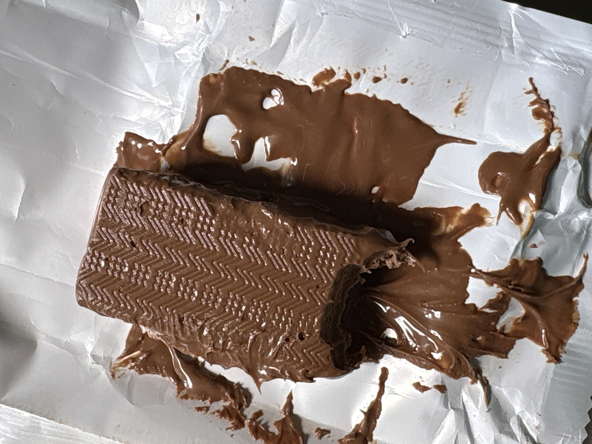 A partially consumed, rectangular chocolate bar sits atop crumpled silver foil packaging. The bar has a ridged, textured surface. Melted chocolate surrounds the bar, pooling on the foil. No people or other objects are visible. The location is unknown.