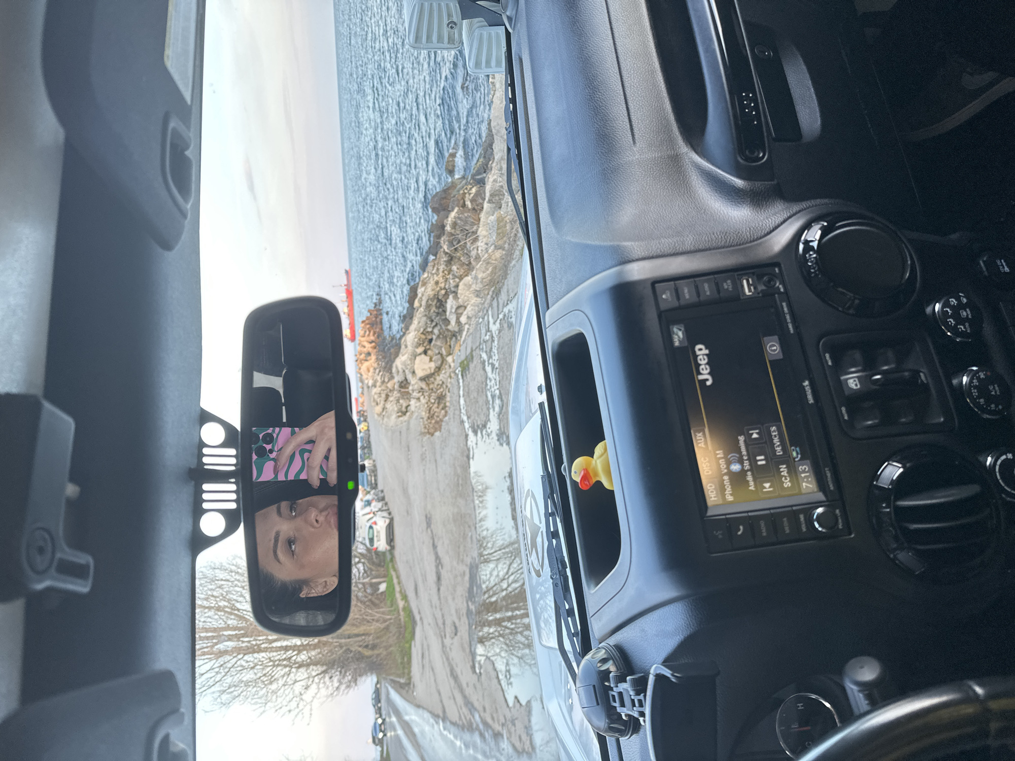 The image shows the interior of a Jeep vehicle. The center console is visible with a Jeep infotainment system displaying audio information and various control buttons. A yellow rubber duck is positioned in the windshield corner. In the rearview mirror, a woman's face is visible, holding a phone with a patterned case. The windshield provides a view of a coastal landscape with a rocky shoreline and the sea. This coastal landscape indicates this is likely in Varna, Bulgaria.