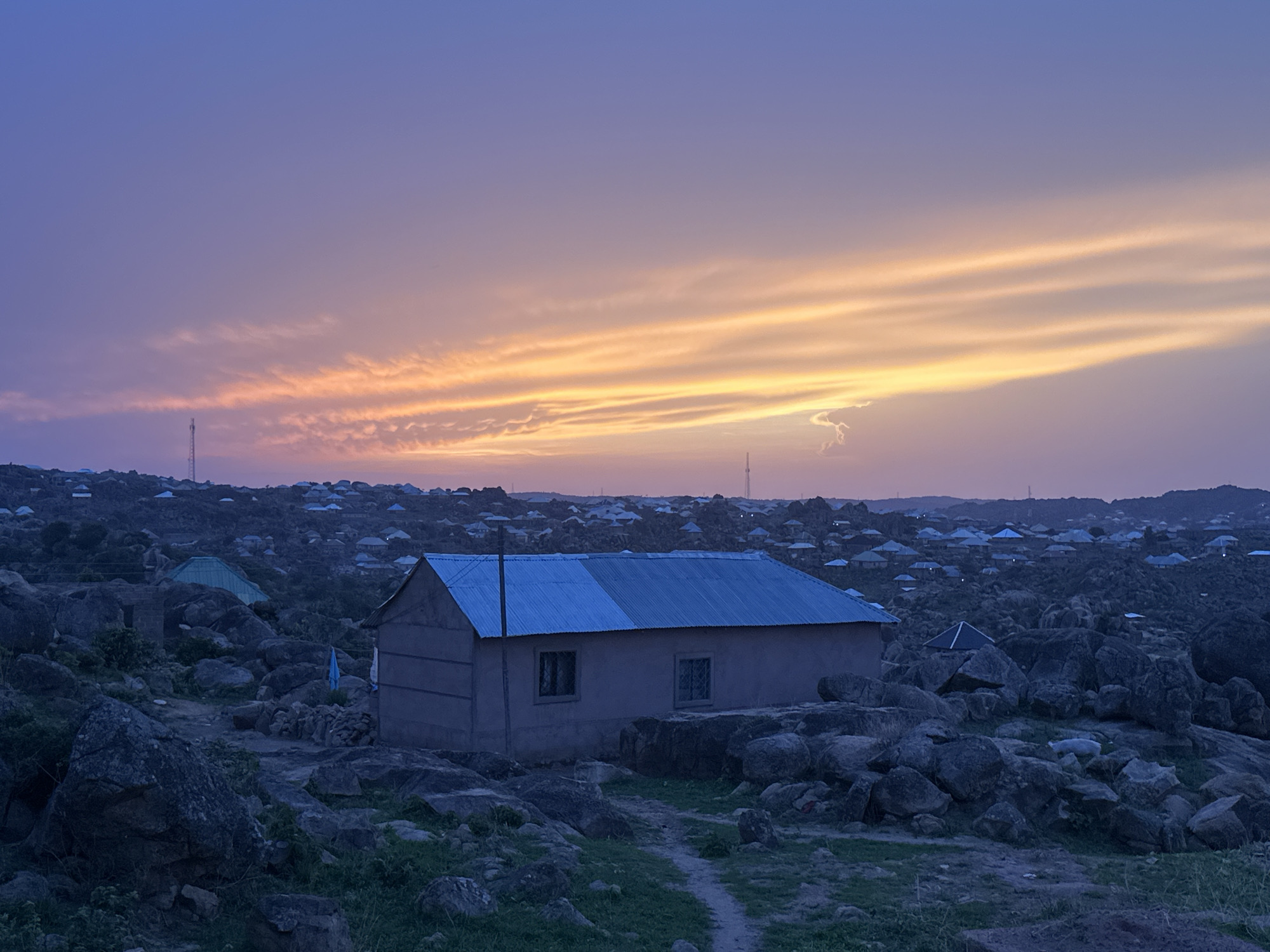 The image depicts a small, rectangular building with a blue corrugated metal roof, located in a rocky, uneven terrain. The building has two visible windows. The ground is covered with large rocks and sparse vegetation. In the background, numerous similar buildings can be seen scattered across the landscape, suggesting a populated area. The sky is streaked with shades of orange and purple, indicating either sunrise or sunset. The environment appears rural or on the outskirts of a city. The location can be further described as Jos, Nigeria, based on the landscape and architectural style.