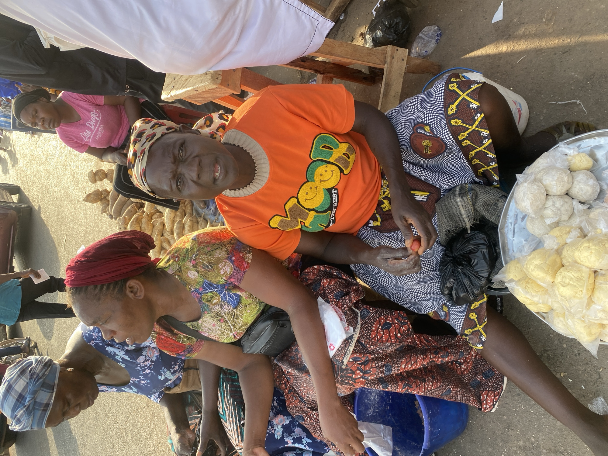 The scene depicts an outdoor street or market setting in Sabon Tasha, Nigeria, under direct sunlight, likely late afternoon or early morning given the light quality and shadows.
In the foreground, three adult women are seated on low surfaces. On the far left, a woman with a blue and white plaid headwrap and a blue floral dress is visible, looking downwards, her hands near a blue plastic bucket. Adjacent to her, a woman wearing a red headwrap and a colorful floral-patterned shirt, with a black cross-body bag, is looking towards her right. In the immediate foreground, facing the viewer, is a woman wearing an orange t-shirt with "MOOD THE BEST" lettering and an animal-print headwrap. She holds a small red fruit in her hands, appearing to peel or prepare it. Directly in front of her is a metal tray containing several small, light-colored, individually plastic-wrapped food items. A black plastic bag is on the ground near her left leg.
In the midground and background, another woman in a pink t-shirt with "San Diego California" text and a black headwrap sits on a red plastic stool, positioned behind piles of unpeeled root vegetables, possibly yams or sweet potatoes, stacked on the ground. A partial view of a red vehicle and other indistinct figures are visible further in the background, indicating a public thoroughfare. A wooden structure is partially visible on the right.
