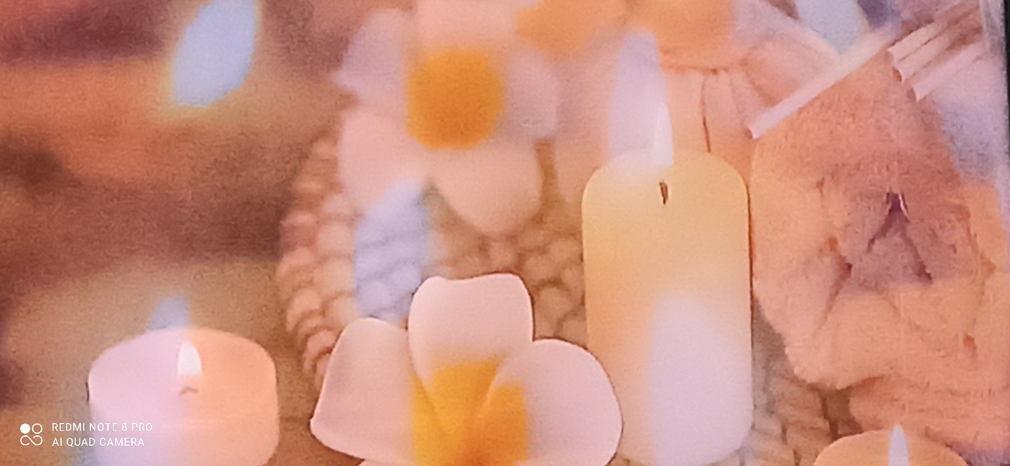 The image shows a close-up of a still life arrangement, likely intended for relaxation or decoration. Two white candles are visible, one in the foreground left and another taller, yellowish candle in the middle right. Both candles appear to be lit, emitting a soft glow.
In front of the candles are two white flowers with yellow centers, resembling frangipani or plumeria. Their petals are delicate and appear soft. Behind the flowers and candles, the background consists of a textured, possibly woven material in warm, neutral tones of beige and light brown. There are also hints of what might be other decorative elements, such as rolled-up fabric or reeds, adding to the serene and spa-like atmosphere.
The image's bottom left corner displays a watermark indicating "REDMI NOTE 8 PRO" and "AI QUAD CAMERA," suggesting the photograph was taken with a smartphone manufactured by Xiaomi.
The prompt mentions the location as Sedati Agung, Indonesia. While the image itself doesn't contain any explicit geographical markers, the presence of the watermark from an Indonesian market phone could subtly support this location context. The scene depicts a common decorative setup found in homes, hotels, or spas, aiming to create a calming ambiance. No people are visible in the image.
