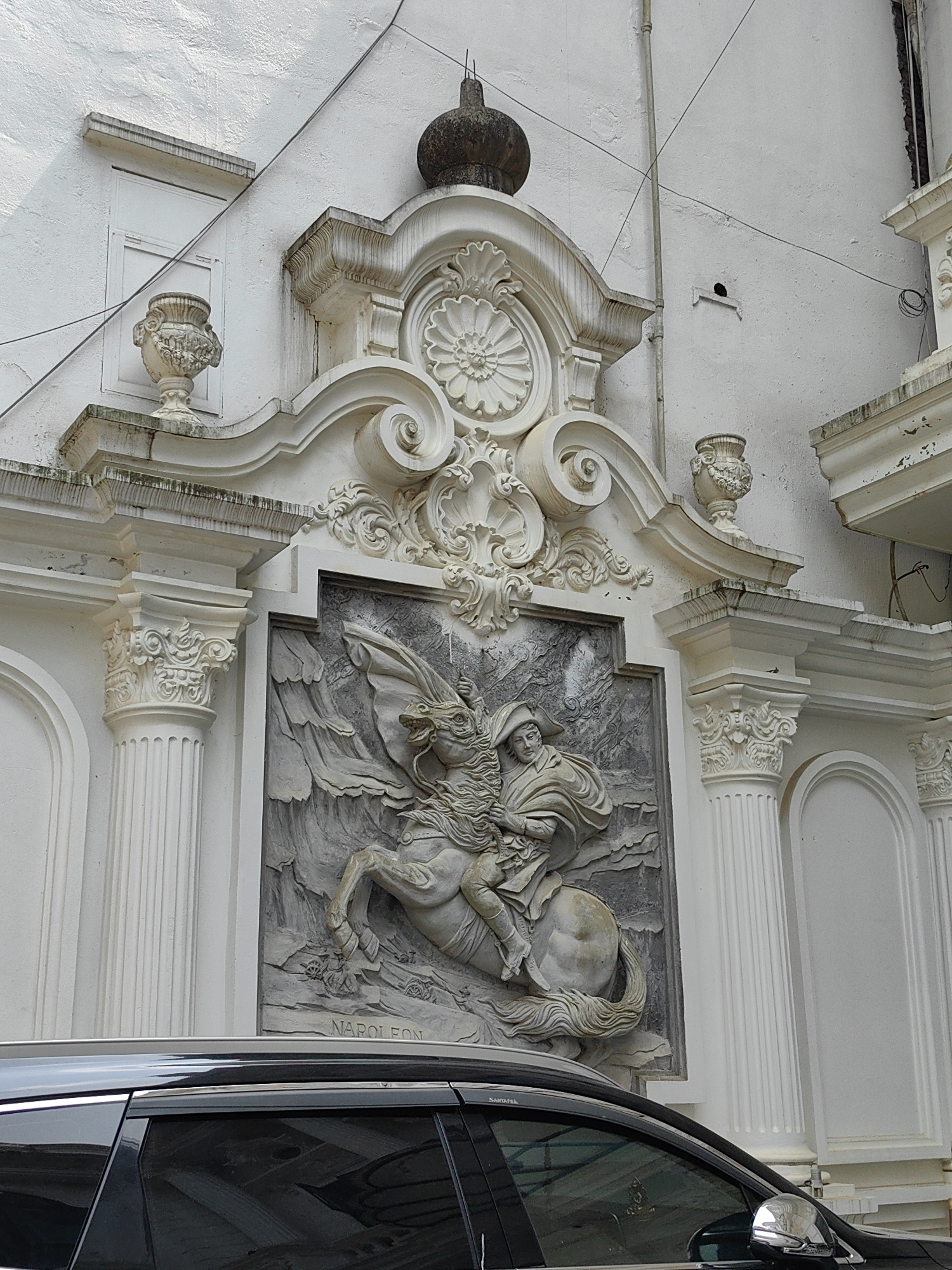 A large, ornate architectural relief carving of Napoleon Bonaparte on horseback is prominently displayed on the exterior of a white, stuccoed building in Sa Pa, Vietnam. The relief, rendered in shades of gray, depicts Napoleon in a bicorne hat and flowing cape, riding a rearing horse amidst a stylized mountainous or cloud-like background. The name "NAPOLEON" is inscribed at the bottom center of the relief panel.
The relief is flanked by white pilasters with Ionic-style capitals and is set within an elaborate baroque-style architectural framework. This framework features scrolled volutes, a central floral medallion, and decorative urns atop pedestals. A dark, rounded finial crowns the entire ornamental ensemble at the building's apex. The white facade exhibits signs of weathering, including dark streaks and some visible grime. Multiple black electrical wires are strung across the building's surface, and a vertical pipe runs along the upper left section.
In the immediate foreground, the top section of a black vehicle, likely an SUV, is partially visible. Its roof rack, darkened windows, and a chrome side mirror are discernible, indicating it is parked directly in front of the building.