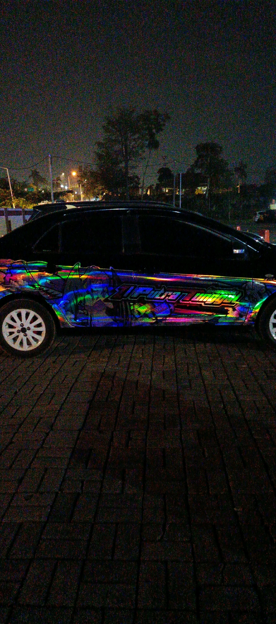 The image captures a black sedan parked at night on a brick-paved surface. The car is adorned with a vibrant, iridescent vinyl wrap featuring colorful abstract patterns and stylized text. The background reveals a dimly lit urban environment with trees, streetlights, and distant buildings. The overall scene suggests the car is parked outdoors in a city setting, possibly a street or a parking lot in Pangkalpinang, Indonesia, given the context provided. The image quality is somewhat grainy due to low light conditions.