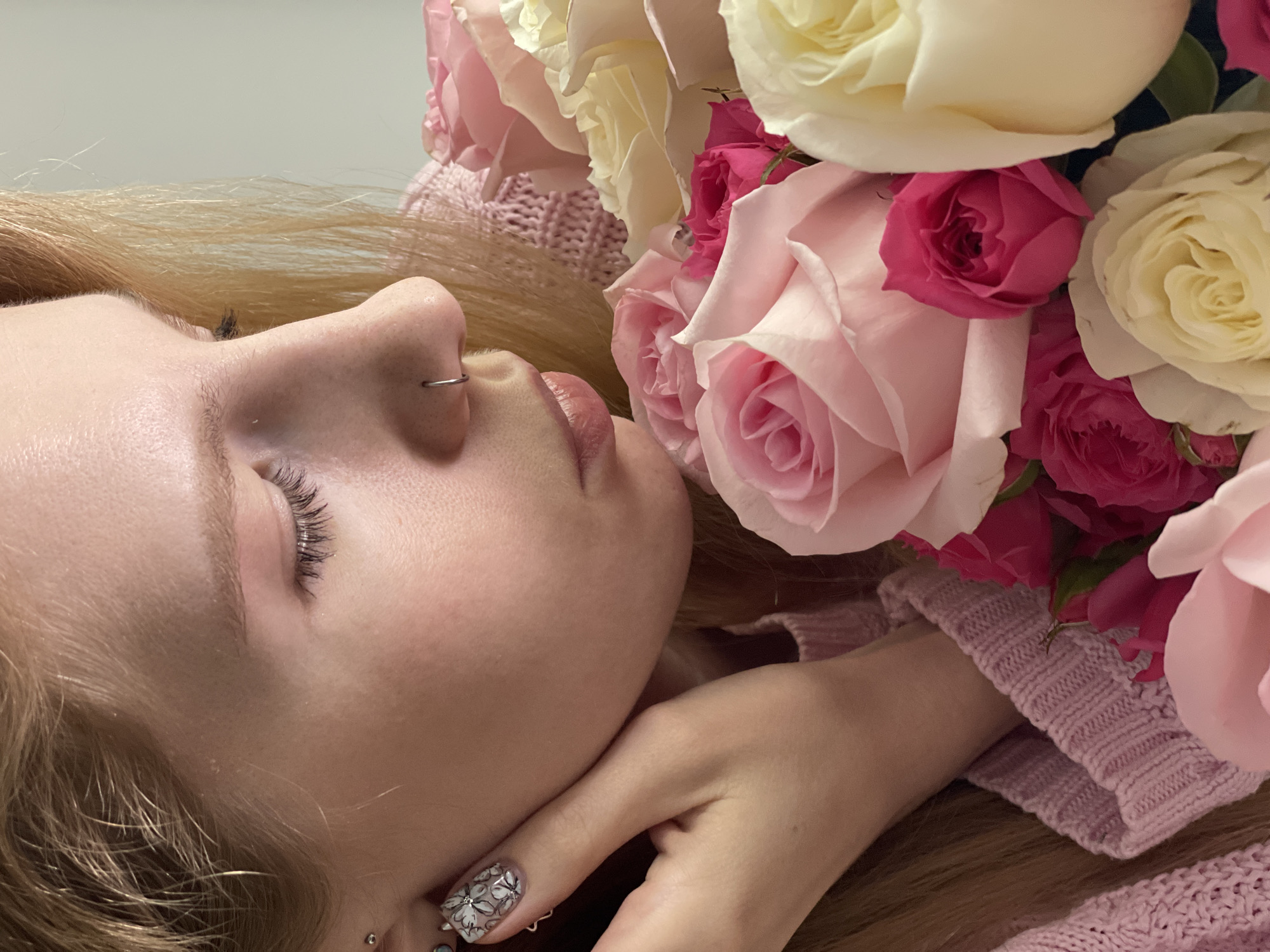 An intimate, close-up shot captures a young woman lying down in profile, her eyes gently closed, suggesting a moment of peace or rest. Her blonde hair spreads out around her, contrasting with her fair skin. She wears a small silver hoop piercing in her nose and a subtle stud earring in her earlobe.
A significant portion of the image is filled with a lavish bouquet of roses, predominantly in soft pinks, vibrant fuchsia, and creamy white hues. These flowers are positioned close to her face and hand, creating a tender and romantic atmosphere. Her left hand is visible, resting near her face, revealing a detailed floral nail art design on her ring finger, featuring white flowers with dark outlines and tiny embellishments.
The woman is partially clad in, or resting upon, a light pink knitted garment, possibly a sweater or blanket, which adds to the soft, comforting aesthetic of the scene. The background is simple and out of focus, appearing as a muted, light-colored surface, emphasizing the subjects. The overall lighting is soft and diffused, enhancing the serene and tranquil mood. There is no visible text in the image. The location context is Izhevsk, Russia, although no specific landmarks or signs within the image indicate this.
