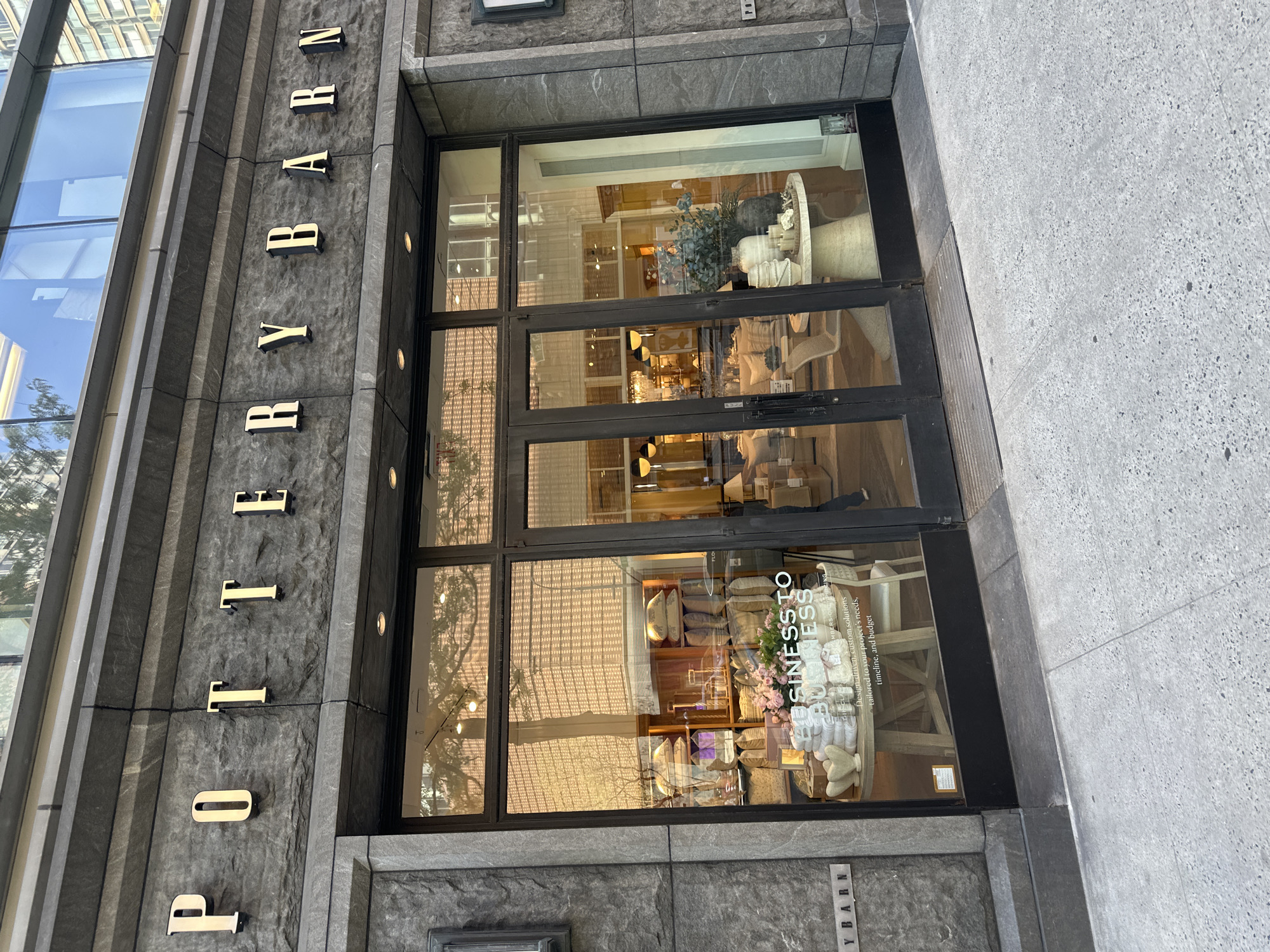 The image shows the entrance to a Pottery Barn store, likely located in a densely populated urban area of New York City. The store's name is prominently displayed in vertical, gold lettering on the exterior wall. The storefront features large glass doors and windows, reflecting the surrounding buildings and sky. A window display showcases home decor items. A sign on the door reads "Business To Business"