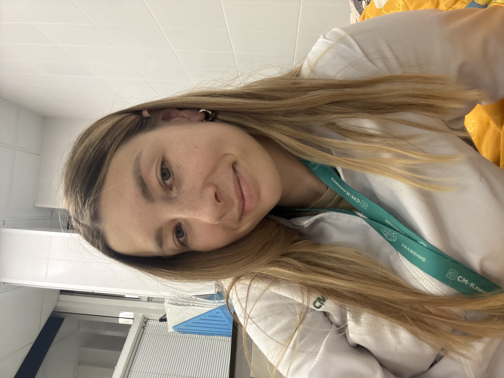 A young woman with long blonde hair and freckles is taking a selfie in a tiled room. She is wearing a white jacket with a teal lanyard around her neck. The lanyard has a logo with text that appears to be in Russian. In the background, there are white tiled walls and a window with blinds. There is also a blue and white object visible through the blinds. The overall impression is a casual indoor setting, possibly a medical facility or office given the lanyard and the tiled walls. The woman is smiling slightly, looking directly at the camera.