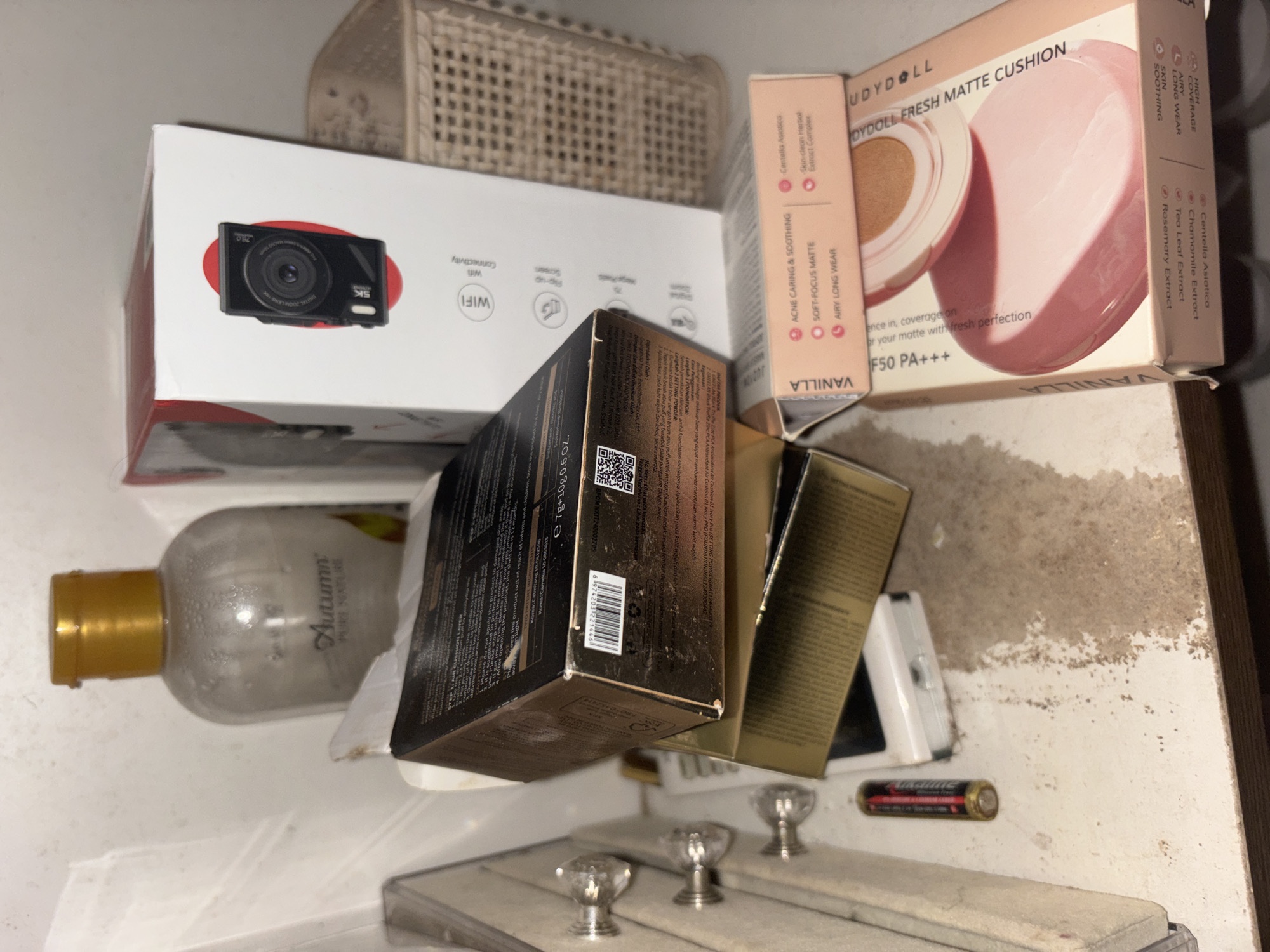 The image depicts a cluttered shelf with various cosmetic products and electronic devices. In the foreground, a pink and white box of "VOYDOLL FRESH MATTE CUSHION" foundation is partially open, revealing a compact. To its left, a golden-brown box is stacked on top of another box of similar color. A white electronic device with a screen is visible beneath the golden boxes, with a single AA battery lying to its left. Behind these items, a clear plastic bottle with a gold cap, labeled "Autumn PURE NATURE," stands upright. To the right of the Autumn bottle, a white box with a red circular emblem featuring a camera lens is prominent. A woven basket is situated behind the white box. The shelf surface appears dusty and stained, suggesting a lack of regular cleaning. The overall environment appears to be an indoor storage area, possibly a shelf in a home or a retail space in North Jakarta, Indonesia. There are no individuals present in the image, and no actions or interactions are depicted.
