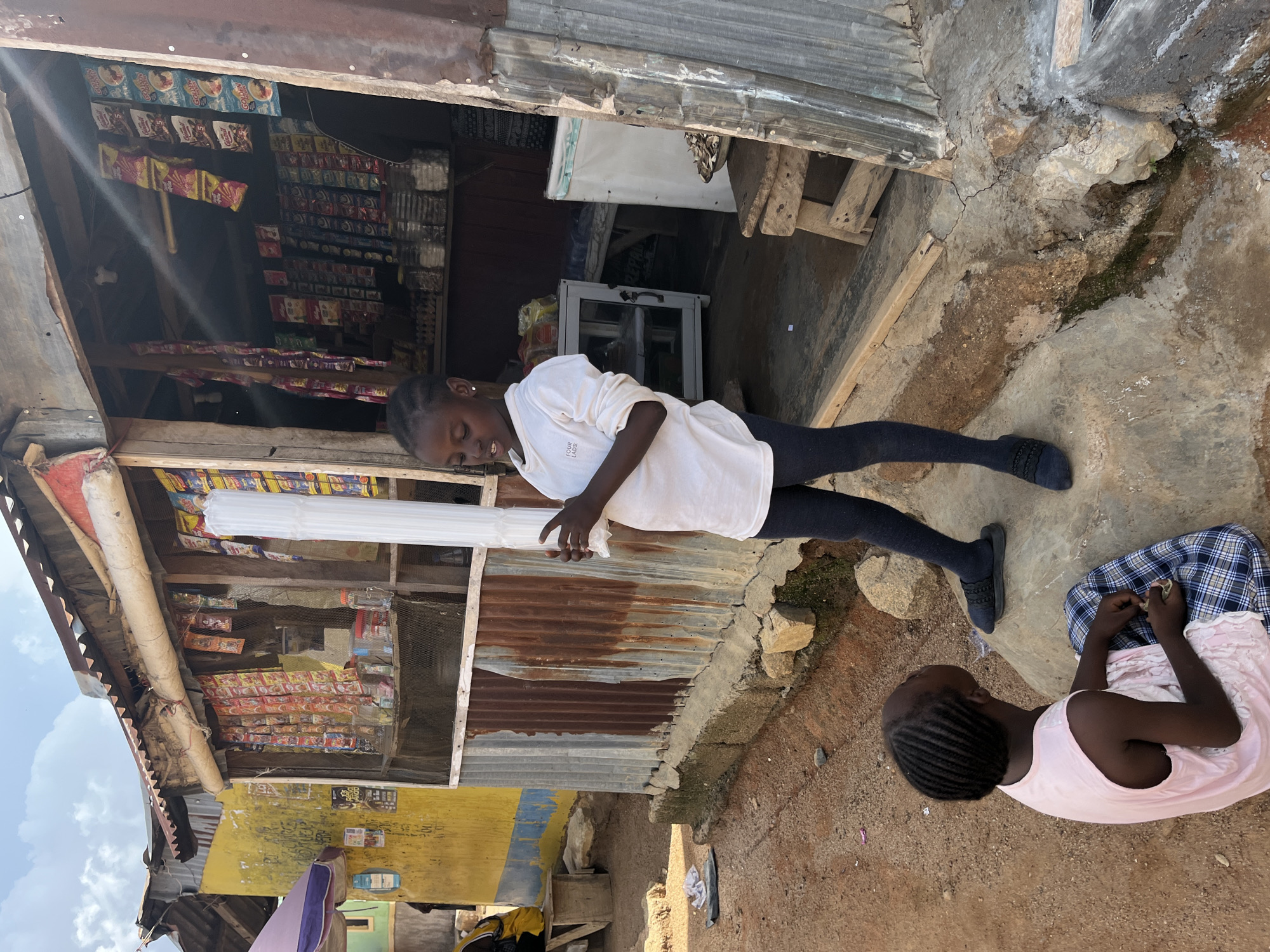 A young Black girl stands holding a long, white, cylindrical object outside a small, open-air shop in Jos, Nigeria. The shop has a corrugated metal exterior and is stocked with various packaged goods. The girl is wearing a white long-sleeved shirt and dark leggings. Behind her, another young Black girl is seated on the ground, looking down at something in her hands. She is wearing a light pink tank top and a blue and white checkered skirt. The ground is dusty and appears to be a dirt road. The environment suggests a developing urban area with simple structures and a sunny day.