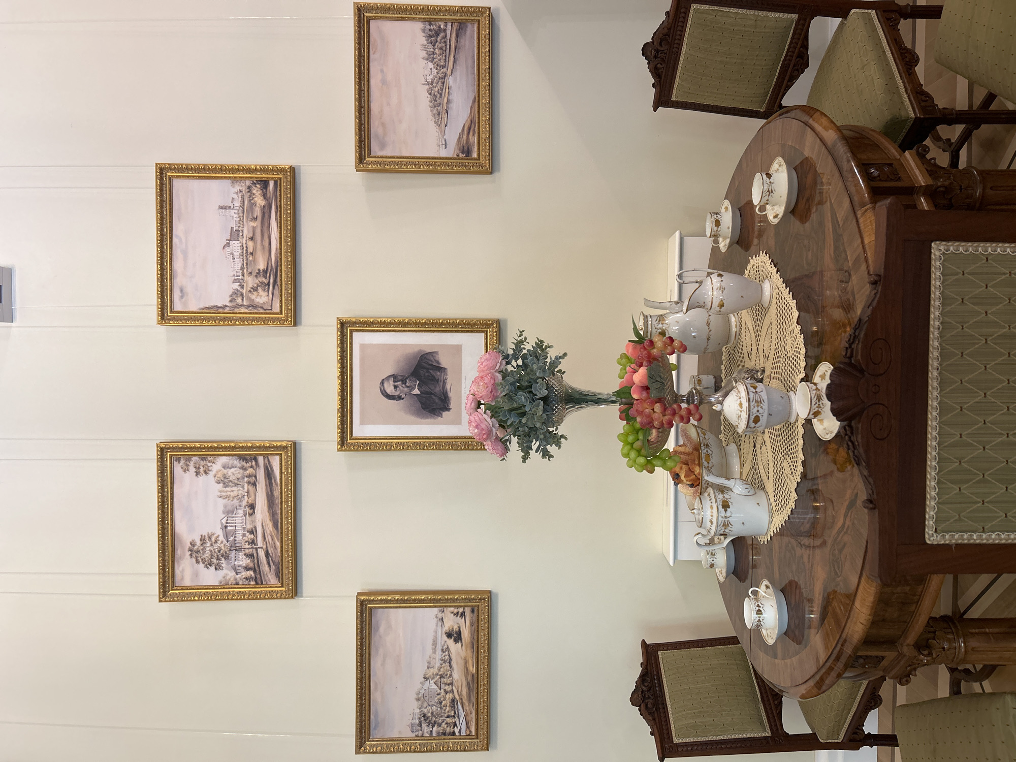 The image depicts a dimly lit, formal dining room, likely within a historical museum or estate in the village of Dobasnyanskoye, Belarus. Five framed landscape paintings adorn the pale yellow walls, arranged in a symmetrical pattern around a central portrait of a distinguished gentleman. The paintings, all in ornate golden frames, appear to be watercolor or oil depictions of rural estates and riverside towns.
In the foreground, a polished, dark wood oval dining table is set for tea. Two ornately carved, upholstered chairs with green fabric seats flank the table. On the table, a white porcelain teapot, sugar bowl, and creamer are arranged on a delicate, cream-colored lace doily. A glass vase filled with a bouquet of pink flowers and greenery sits behind the tea set, and a fruit bowl overflows with grapes and peaches. The table also holds three teacups and saucers, each adorned with delicate floral patterns.
The overall ambiance is one of refined elegance and quiet repose, suggesting a preserved moment in time. The lighting is soft, casting subtle shadows and highlighting the rich textures of the wood and fabrics. There are no people visible in the scene.