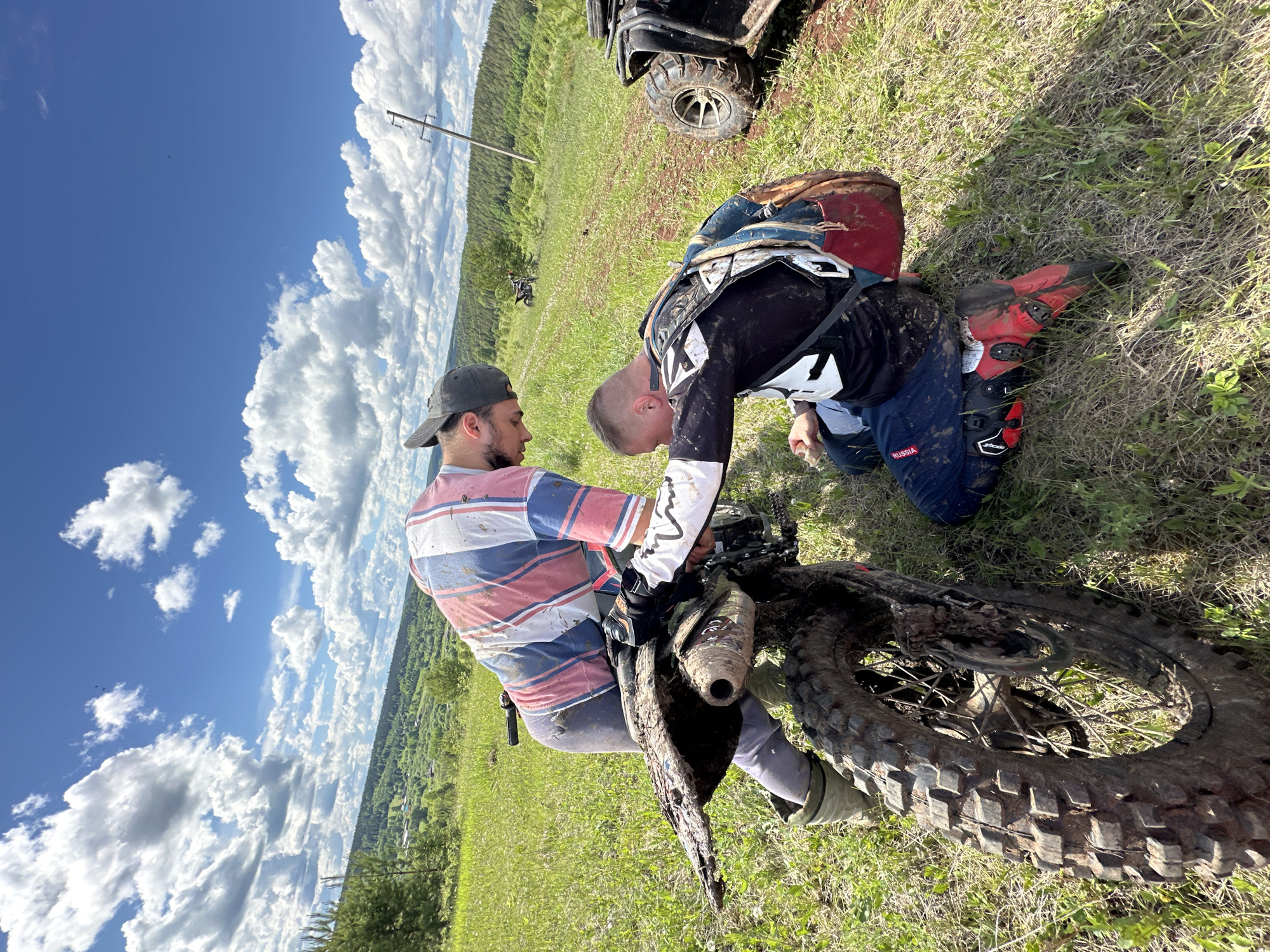 The image depicts two men with a dirt bike in a grassy field under a partly cloudy sky, possibly in the vicinity of Ilyinsky, Russia. One man, wearing a striped shirt and a baseball cap, is seated on the dirt bike. The other man, dressed in motocross gear, is kneeling beside the bike. Both individuals and the bike are covered in mud. An all-terrain vehicle is partially visible in the background. The landscape features rolling hills with forested areas. A power line is also visible in the distance.