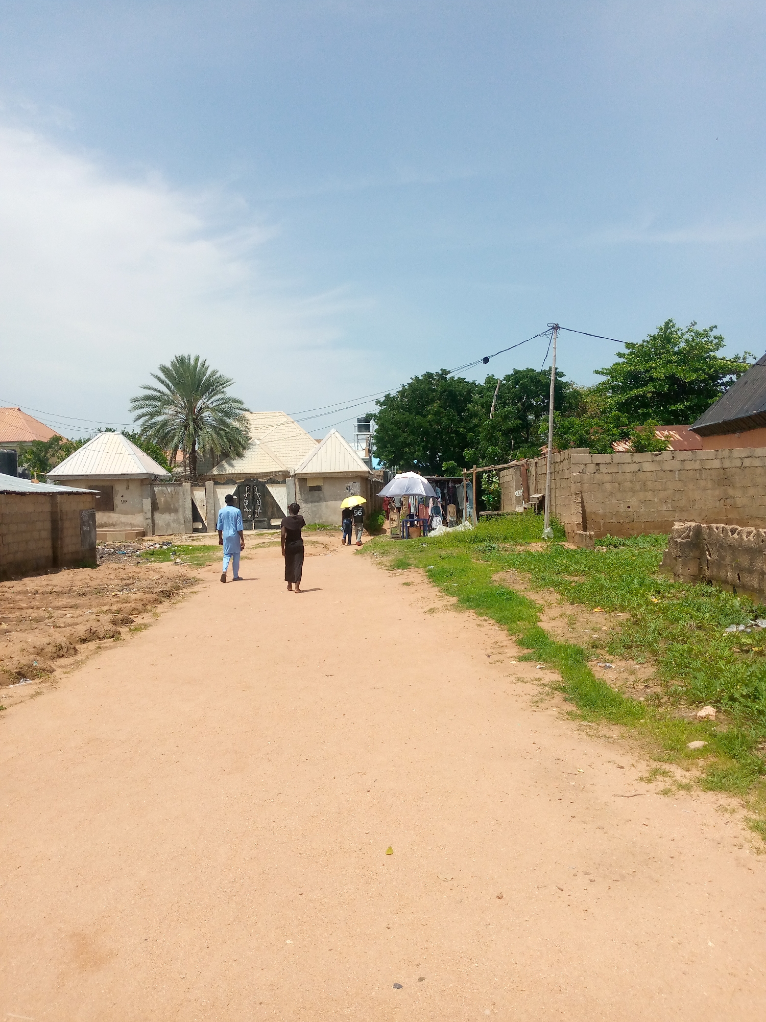 The image depicts a dusty, unpaved road in Jalingo, Nigeria, under a partly cloudy sky. On the left side of the road, a simple cinder block wall marks the boundary of a property with a few structures, including one with a conical roof. A date palm tree stands behind these structures. Further down the road, two individuals, one in a blue traditional garment and the other in a black dress, walk away from the camera.
On the right side of the road, a more substantial cinder block wall runs alongside the path. Beyond this wall, there are more buildings, including a dark-tiled roof. A small market stall is visible with umbrellas providing shade and clothing items displayed. A few people are gathered around the stall. The overall environment suggests a residential or mixed-use area in Jalingo, characterized by its unpaved roads and building materials. Power lines are visible strung between poles along the right side of the road.