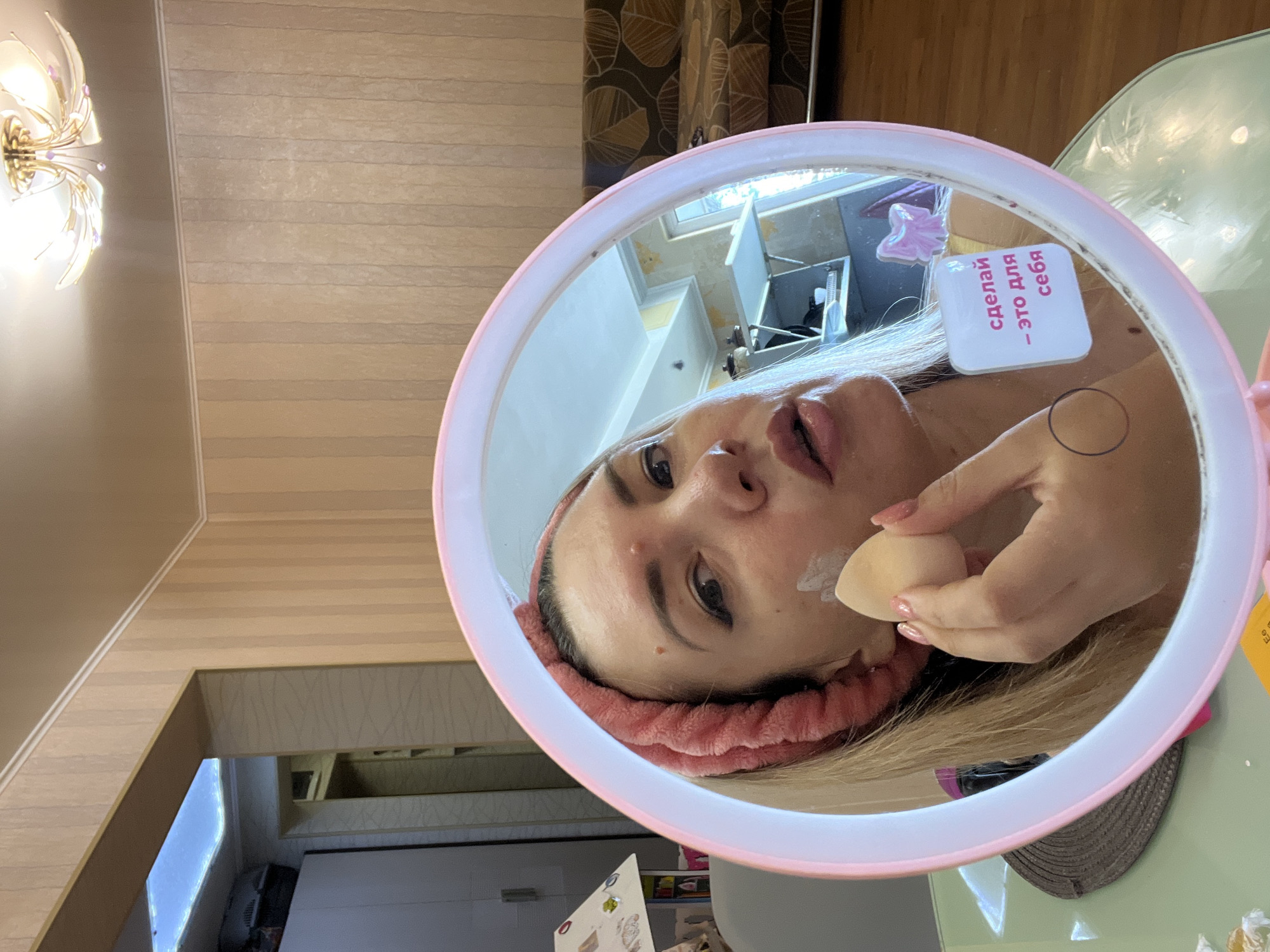 This image shows a woman applying makeup in a bathroom, reflected in a circular mirror with a pink light. She is wearing a pink headband and has blonde hair. A makeup sponge is visible in her right hand, and she is applying a white substance to her cheek. There is a sticker on the mirror with Russian text that reads "сделай - это для себя" which translates to "do it for yourself." The background shows a room with striped wallpaper, a couch, and a window. The location is not specified as Sochi, Russia.