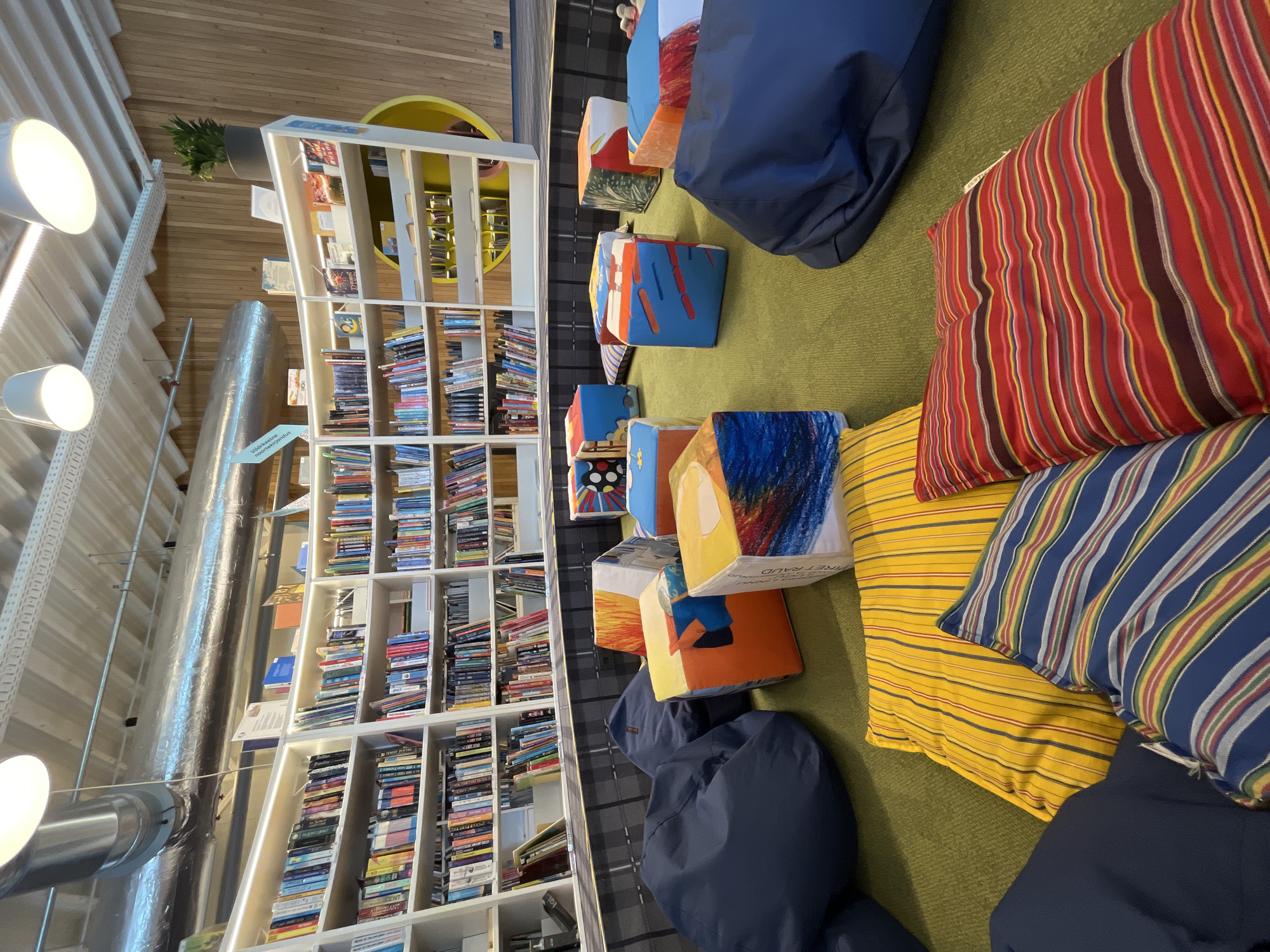 This image depicts a reading nook within a library in Haabneeme alevik, Estonia. The scene features a semi-circular bookshelf filled with numerous books, suggesting a well-stocked collection. In front of the bookshelf, scattered on a green carpet, are several large, colorful, cube-shaped seating elements with abstract artwork and various beanbag chairs. The seating area appears designed for comfort and relaxation, suitable for individuals to read or study. The environment is lit by overhead lights and the overall aesthetic suggests a modern and welcoming library space. There are no people visible in the image.