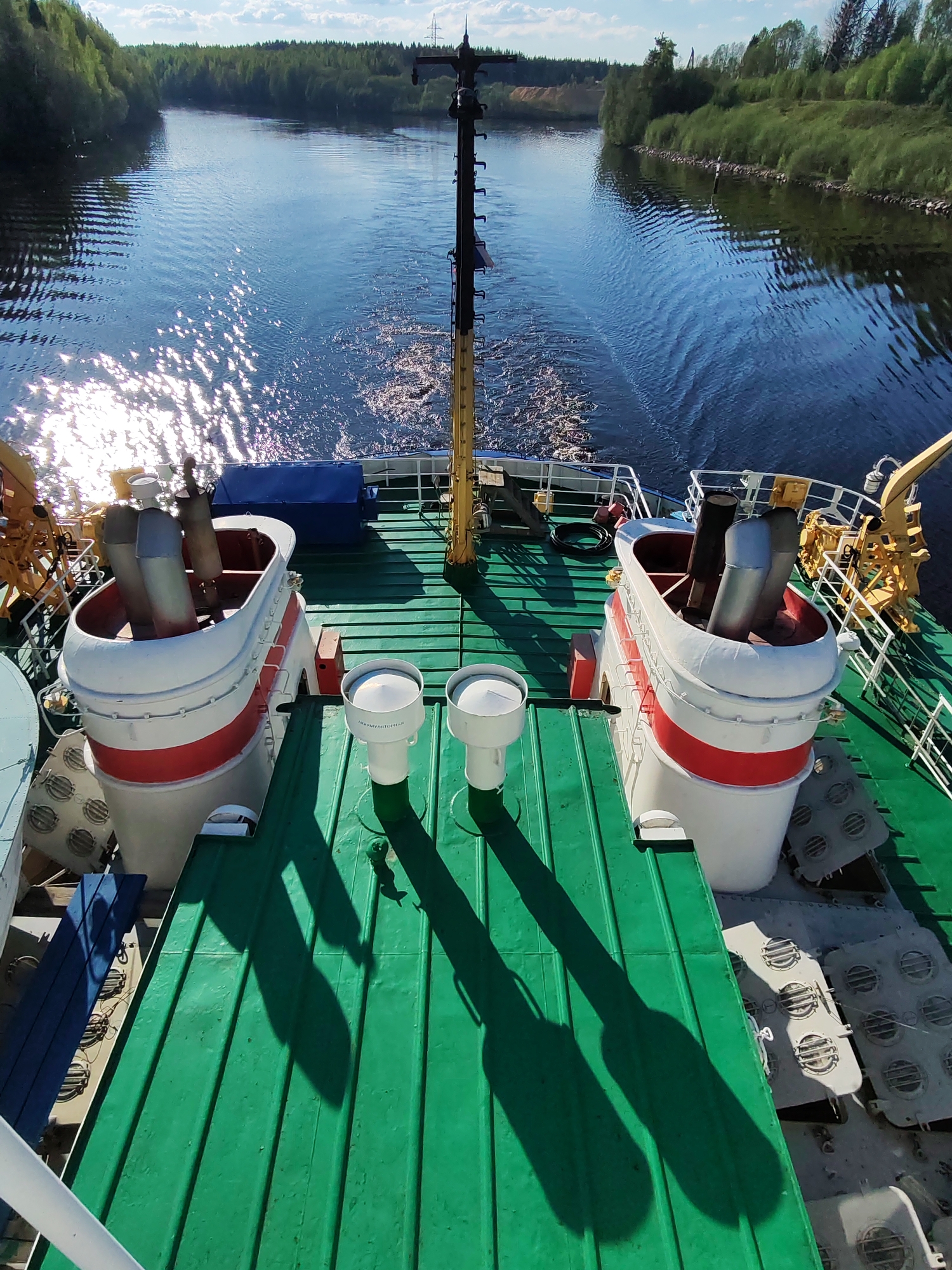 A view from the deck of a boat moving along a canal in Depo, Russia on a sunny day. The canal is lined with green trees and the water is reflecting sunlight. The deck is green and has various pieces of equipment, including two large, white and red structures with exhaust pipes, two white cylindrical objects, and other machinery.