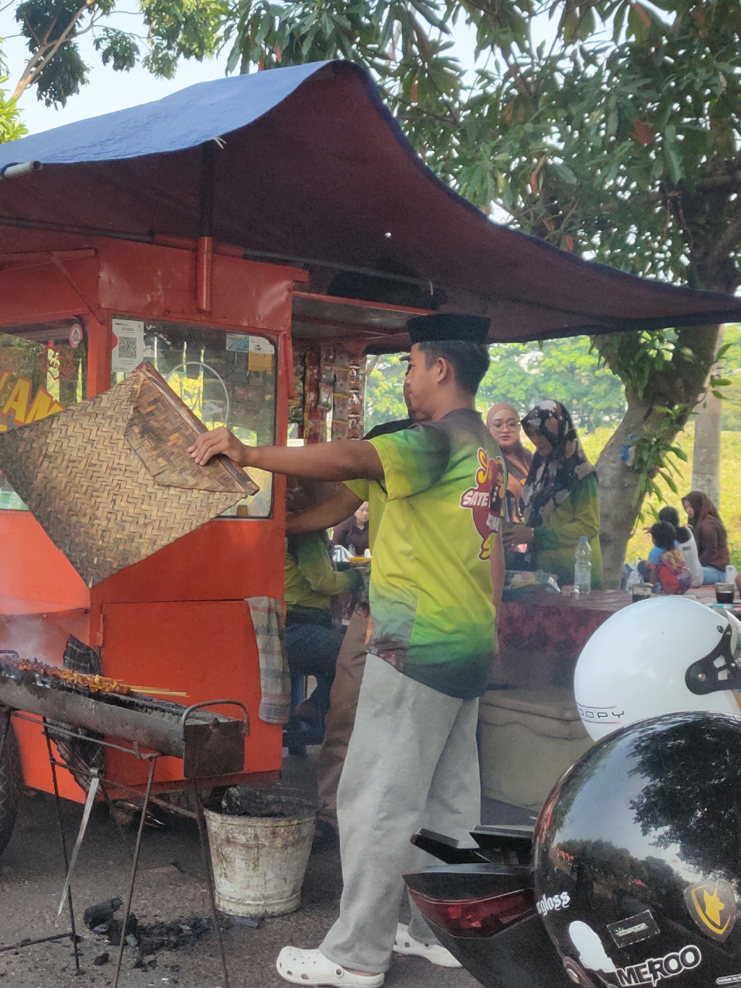 The image depicts an outdoor scene in Ngasem, Indonesia, likely near a street or market. A man wearing a black peci hat and a lime-green t-shirt is standing by a bright orange food cart. He is holding a woven bamboo tray in his left hand and appears to be tending to food being grilled on skewers on the cart. Behind him, two women in headscarves are seated at a table, with one woman looking towards the camera. Other people, including children, are visible in the background, some sitting and some standing. The food cart is covered by a dark-colored tarp, and a grill with skewers of meat is positioned in front of it. A white bucket is placed on the ground next to the grill. In the foreground, two motorcycle helmets are visible, one white and one black, with the black helmet having stickers on it. Trees and foliage frame the background, suggesting a somewhat rural or suburban setting within Ngasem. The overall atmosphere appears casual and suggests a food vendor operation.