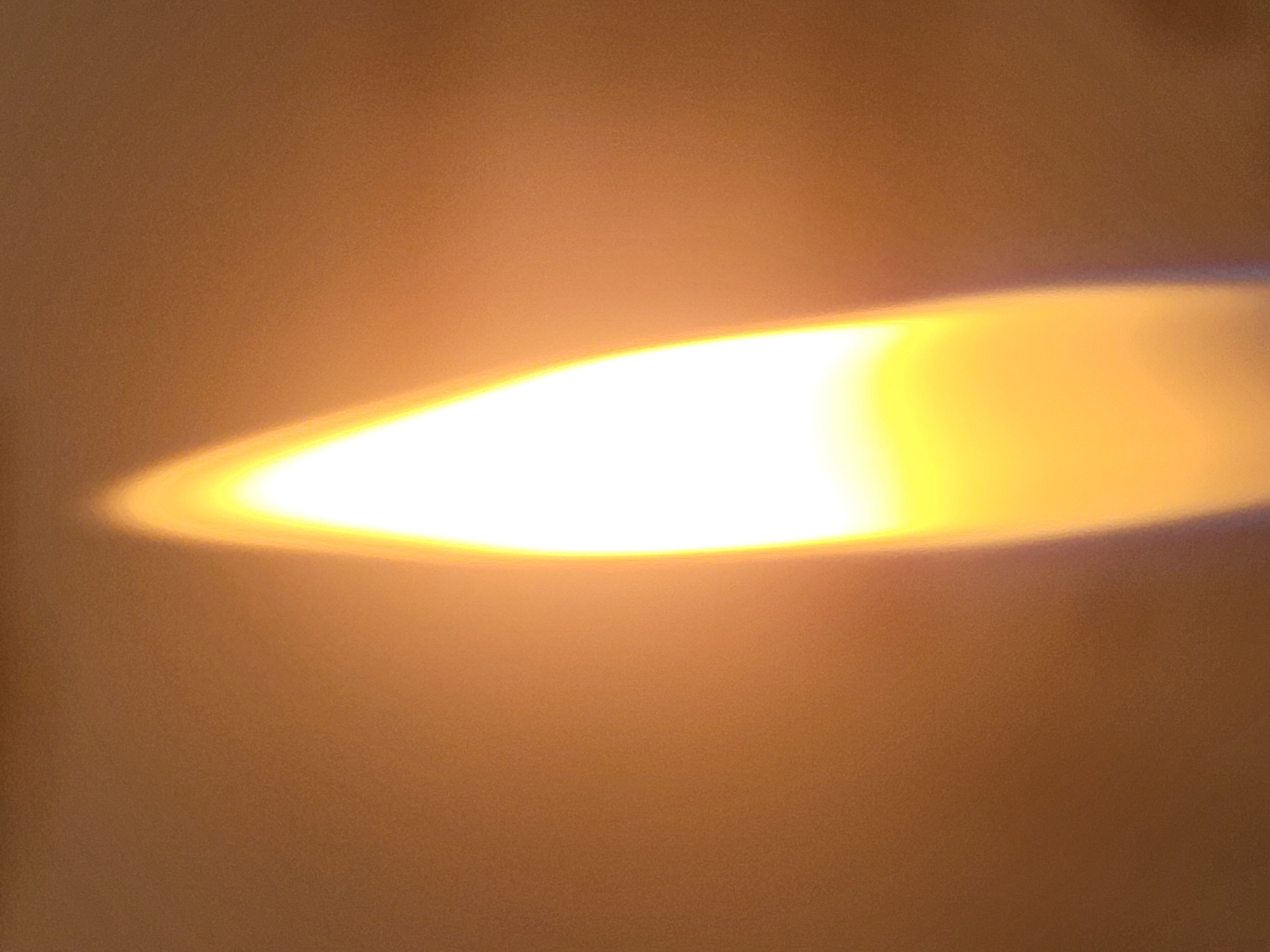 The image shows an out-of-focus, elongated light source with a bright white center fading into a warm yellow-orange towards the edges. The surrounding area is a blurry, light brown-beige. There are no discernible people, subjects, objects, actions, or interactions visible. The environment is indistinct and lacks recognizable features to determine the location, particularly Yerevan, Armenia.