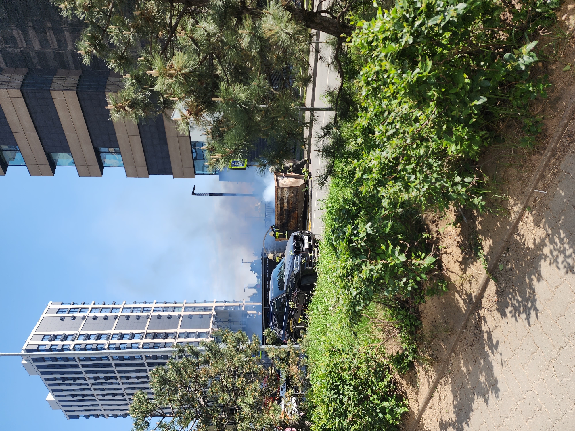 The image shows an incident in Kazan, Russia involving a burning dumpster and a black BMW X5. Firefighters are present, working to extinguish the fire in the dumpster. Smoke rises from the dumpster, partially obscuring the view of the buildings in the background. The setting appears to be a street next to a modern building, with trees and bushes nearby. A pedestrian crossing sign is visible, and a sidewalk is present in the foreground.