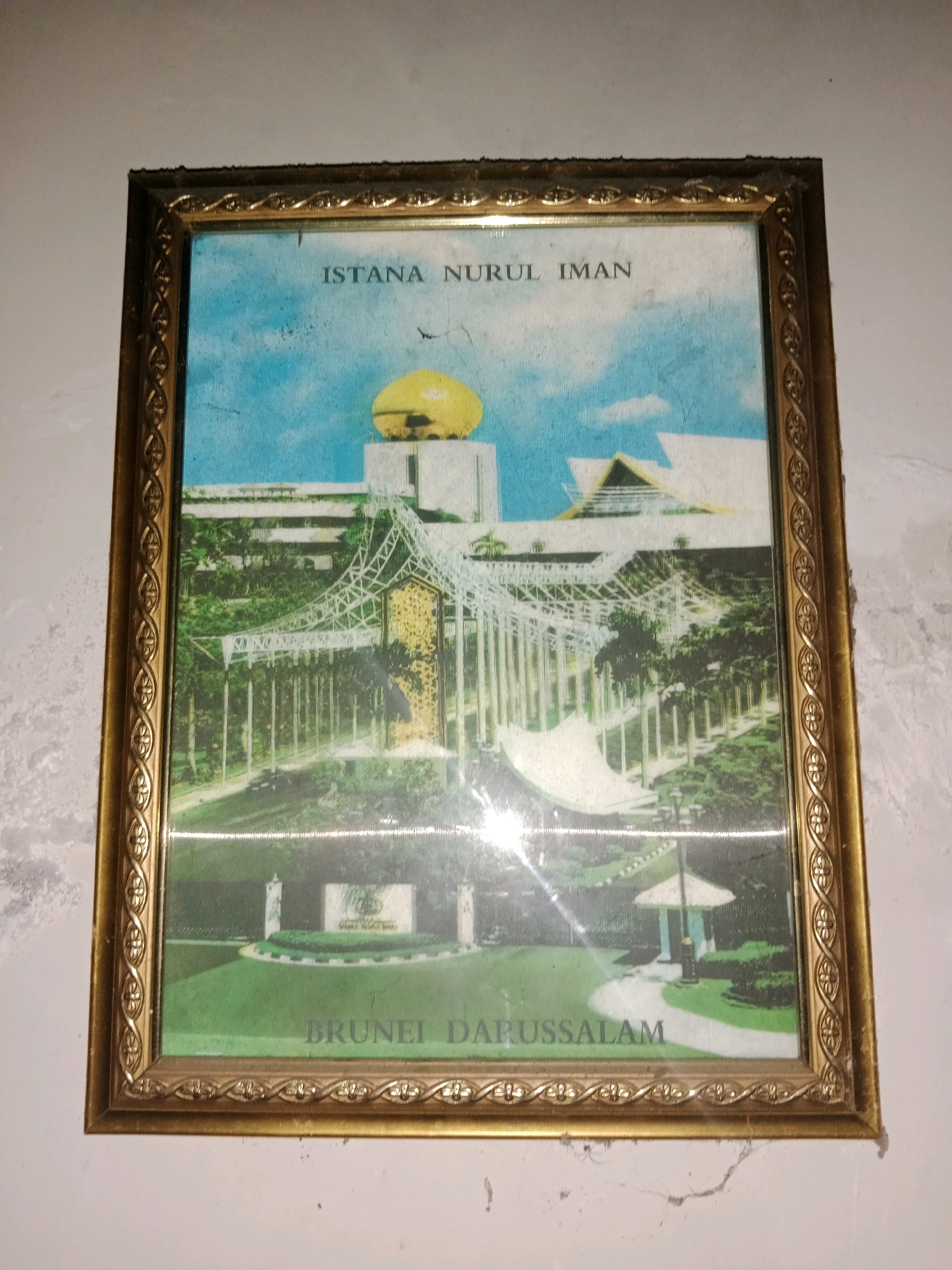The image shows a framed picture of a large building complex with a golden dome, set against a bright blue sky. The building appears to be a palace or government building, with intricate architectural details. There are also trees and manicured gardens surrounding the structure. Text on the picture reads "ISTANA NURUL IMAN" and "BRUNEI DARUSSALAM." The frame is golden with an ornate pattern. The picture is hanging on a textured, white wall, which shows some signs of wear and tear.
