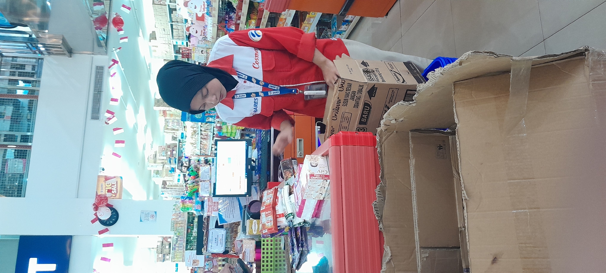 A woman wearing a red and white uniform with a black hijab stands behind a checkout counter in what appears to be a retail store in Rogojampi, Indonesia. The woman is looking down and handling a box. In front of her on the counter are various products, including boxes of soap. Behind the counter, a computer monitor displays a screen, and shelves stocked with merchandise are visible in the background. A large, open cardboard box is in the foreground, partially obscuring the lower part of the scene. Red and white flags are strung across the upper part of the store. The overall impression is of a typical retail environment with a cashier attending to merchandise.