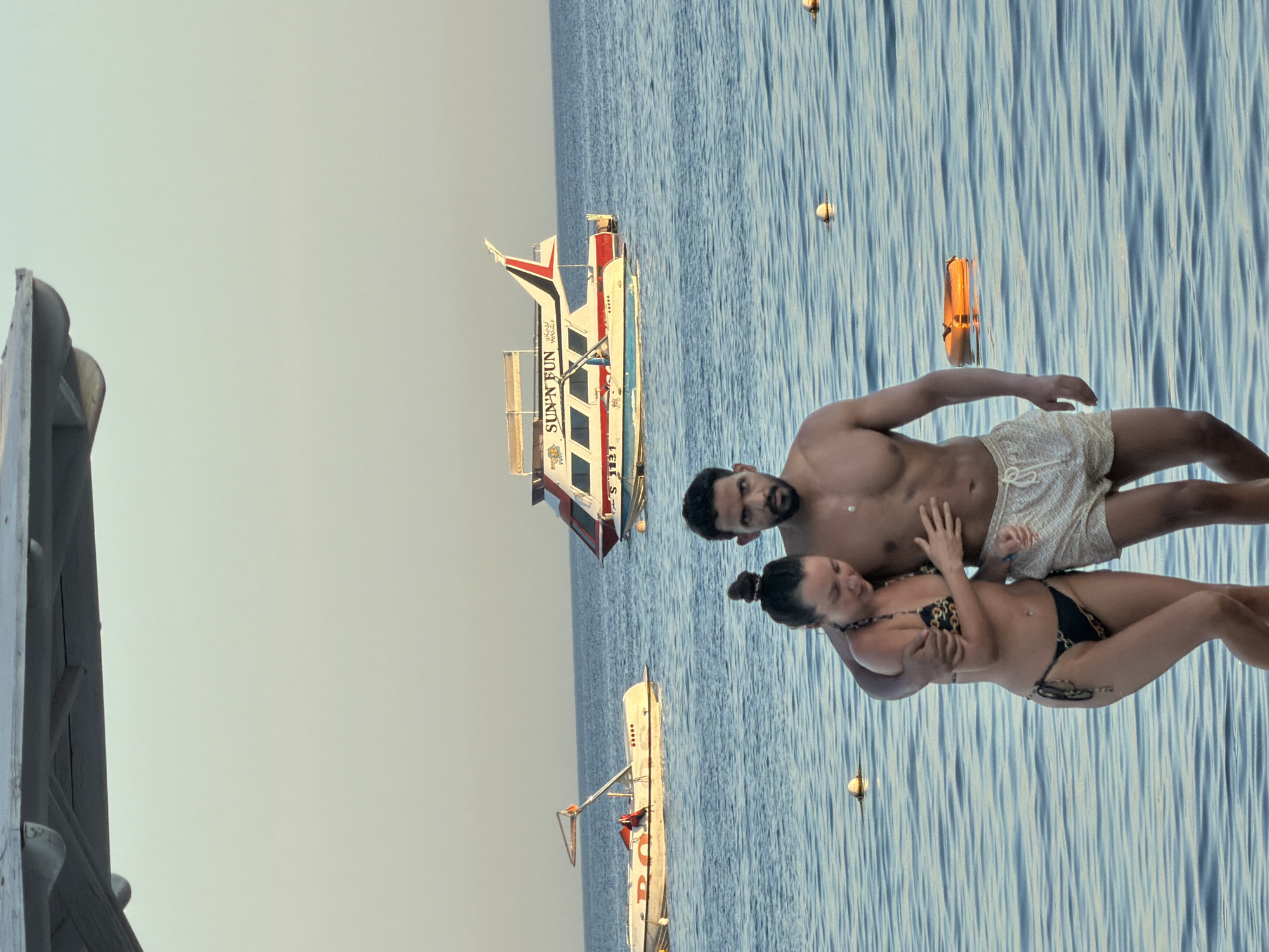 The image depicts a couple walking through shallow water at a beach, likely in Sharm El Sheikh, Egypt. The woman, wearing a black and gold patterned bikini, has her left arm around the man's chest and her right hand resting on his arm. The man, wearing light-colored patterned shorts, has his right arm around the woman's waist. They are walking towards the viewer.
In the background, several boats are visible on the calm blue water. To the left, a white boat with red lettering that appears to say "RO" is partially visible. Further back and to the right, a larger red and white boat with "SUN'N FUN" written on its side is present. The sky is a pale blue, suggesting it is either early morning or late afternoon. The water surface is rippled, and several buoys are scattered throughout the scene. The top of a shaded structure, possibly part of a beach umbrella or canopy, is visible in the upper portion of the frame.