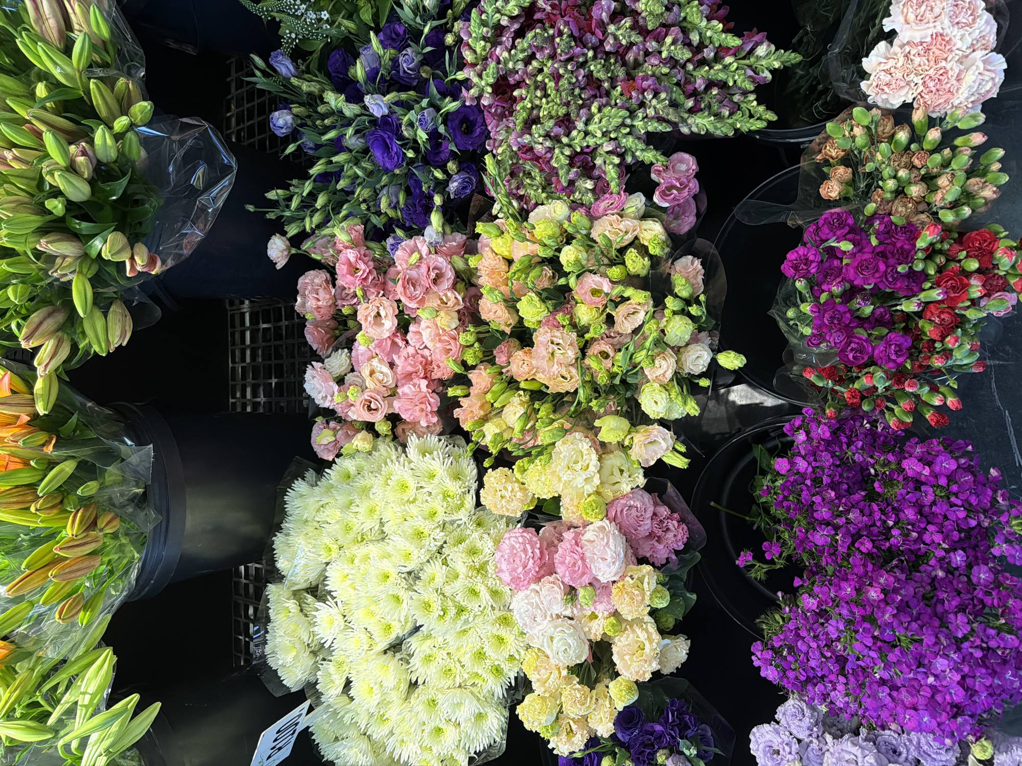 The image shows a variety of flowers displayed in buckets, likely at a florist shop. The flowers include lilies, lisianthus, snapdragons, chrysanthemums, and carnations in a range of colors including purple, pink, white, yellow, and orange. The buckets are placed on a dark surface, and some of the flowers are wrapped in plastic. The photo was not taken in Cape Town, South Africa.