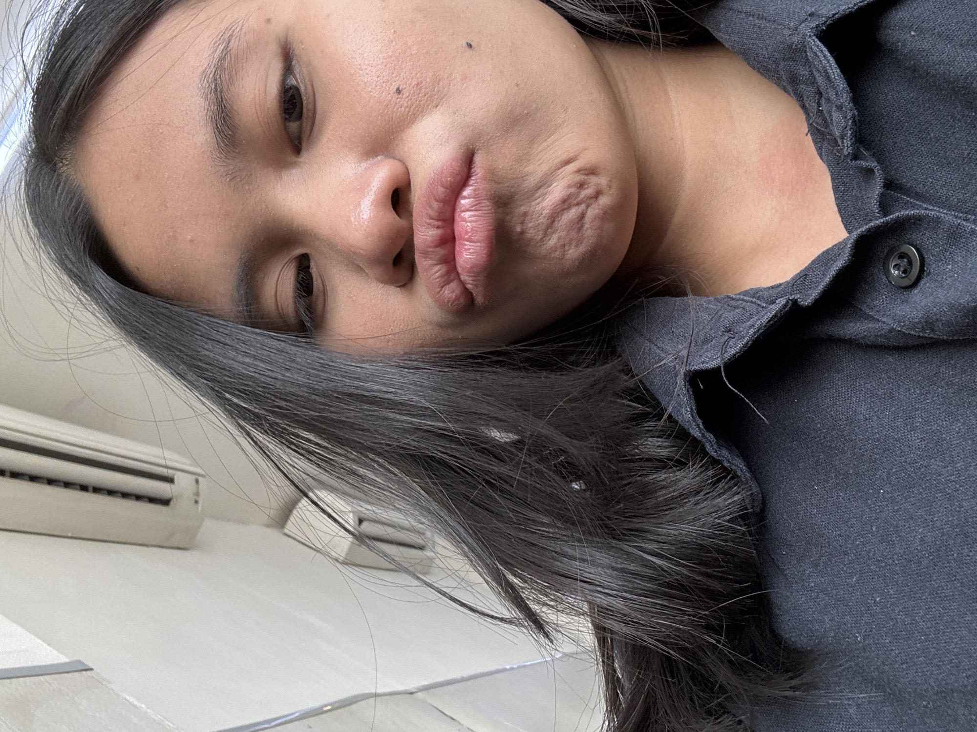 A close-up, low-angle selfie of a young East Asian woman with dark hair and olive skin, taken outdoors in East Jakarta, Indonesia. She is wearing a dark blue collared shirt. Her lips are pursed in a pout, and her chin is wrinkled. Her hair is long and black, partially obscuring her face. The background shows a light-colored wall and what appears to be an air conditioning unit. The lighting is bright, suggesting it is daytime.