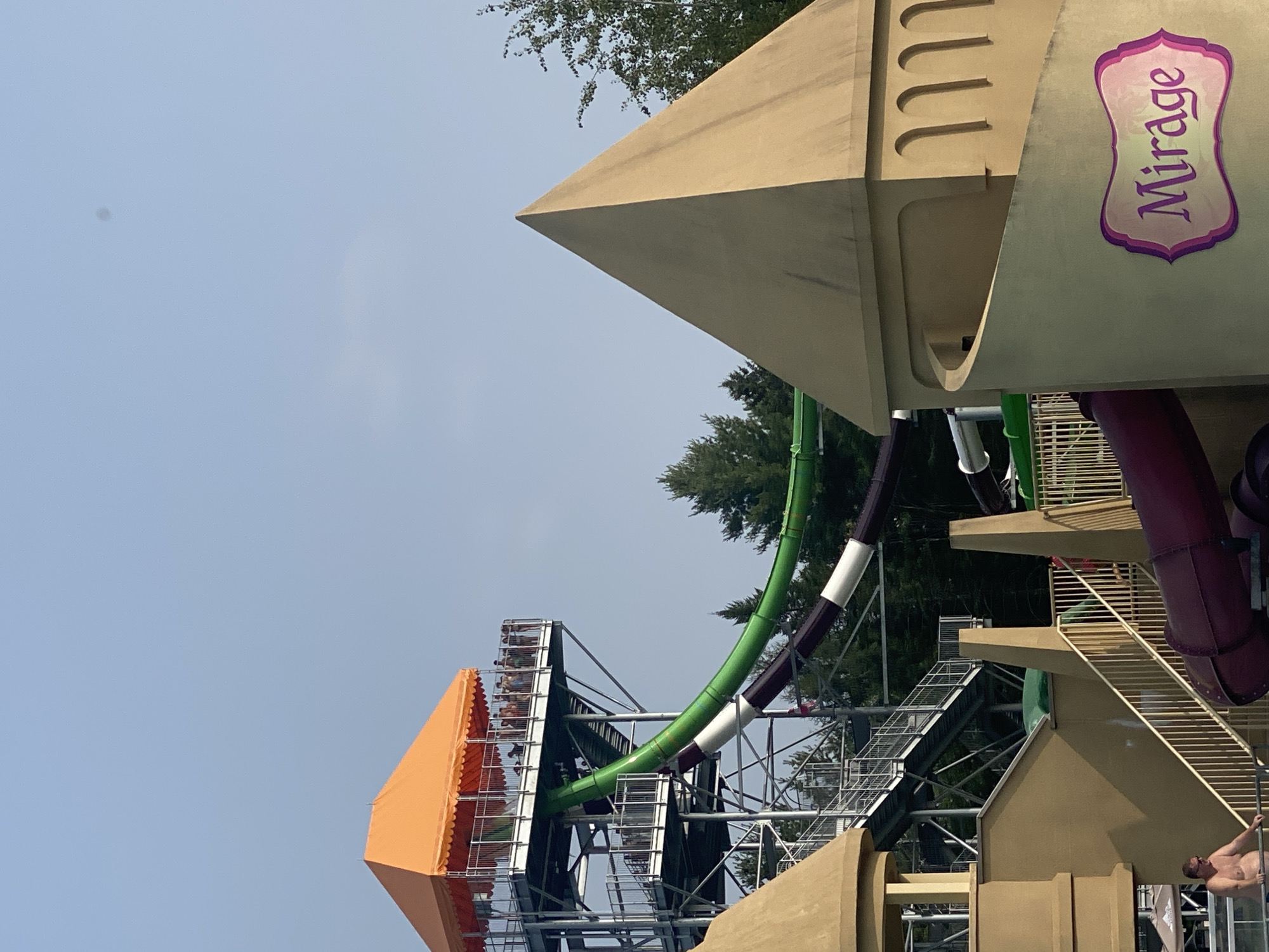 The image captures a section of the Calypso water park in Saint-Gabriel-de-Valcartier, Canada. In the foreground, a tan-colored structure with arched indentations and a curved wall dominates the right side, featuring a decorative plaque with the word "Mirage" in purple lettering. To the left, a tall water slide tower with an orange canopy stands prominently, with multiple levels of stairs and railings. Various sections of a water slide, colored green, purple, and white, descend from the tower. Several people are visible on the observation deck at the top of the tower and one man is seen near a staircase on the left, appearing to be exiting the water. In the background, dense green trees and a clear blue sky form the natural environment surrounding the man-made structures.