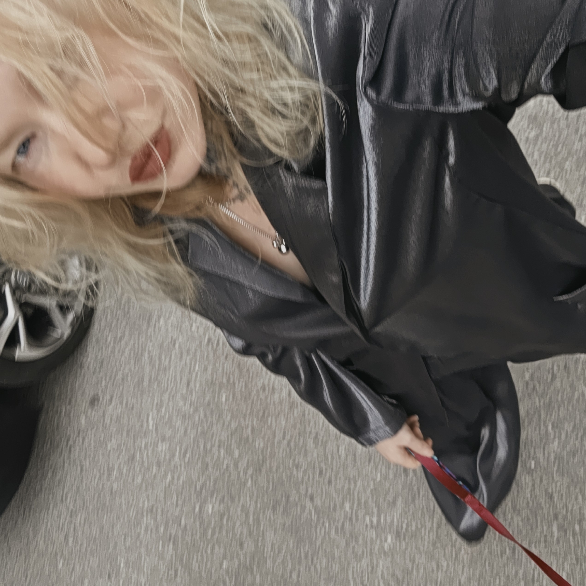 A woman with blonde hair, red lipstick, and visible tattoos on her chest and neck is walking a dog on a leash. She is wearing a shiny, dark jacket and a necklace. She is holding the red leash in her right hand. Part of a car wheel is visible on the left. The setting appears to be an urban street. The city is Saint Petersburg, Russia.