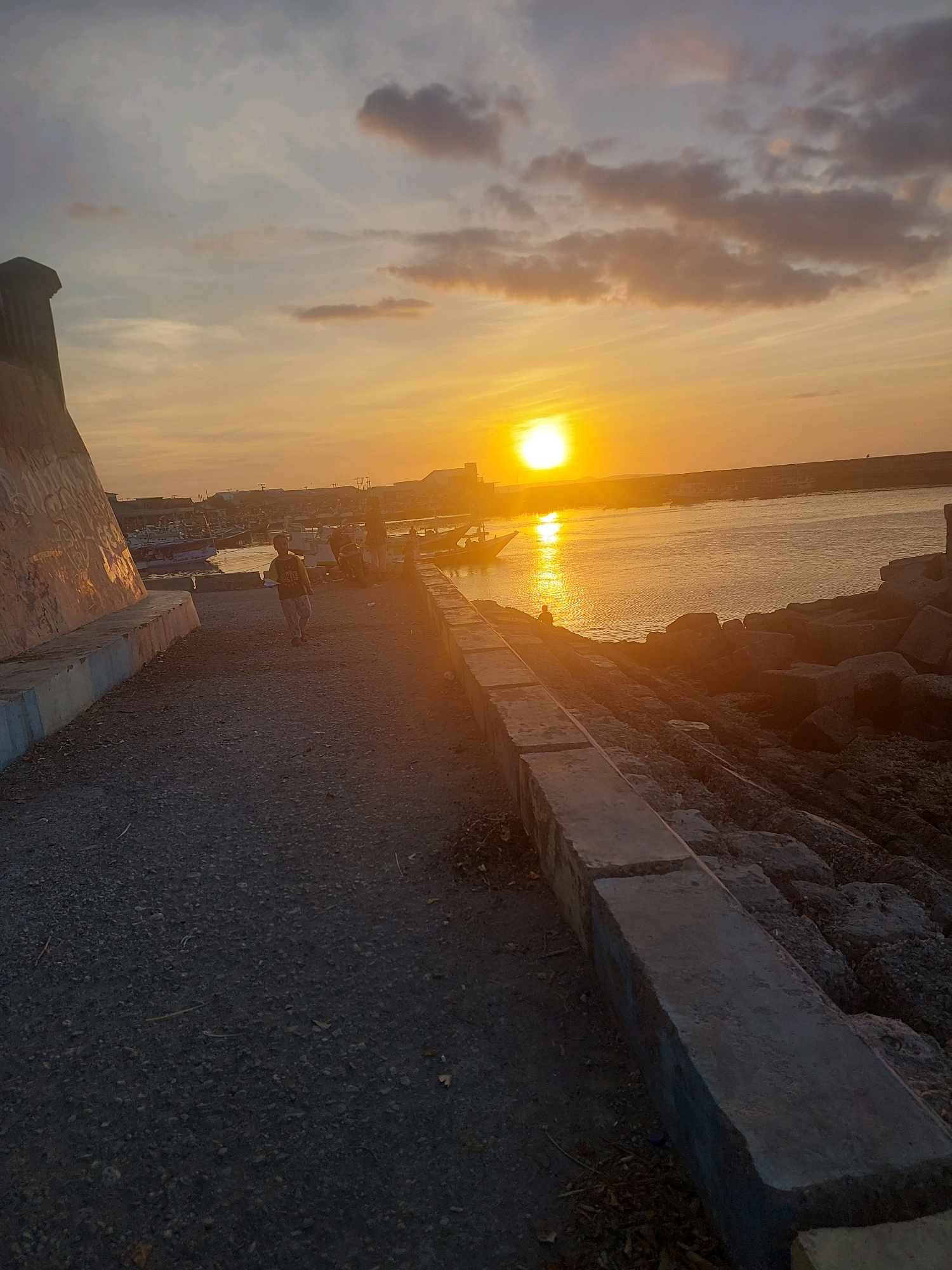 The image depicts a sunset scene at a waterfront in Kupang, Indonesia. The sky is illuminated by the setting sun, casting a warm orange glow across the water and the surrounding environment. In the foreground, a gravel path runs alongside a concrete barrier, leading towards the water. To the left, a large, textured concrete structure with graffiti is visible. A young person in a green t-shirt and patterned pants is walking along the path, facing away from the camera. Further back, a harbor is filled with numerous boats docked or anchored. The water is calm, reflecting the colors of the sky. On the right side of the image, large rocks form a breakwater, with concrete structures also present. In the distance, buildings and more boats are visible, suggesting a populated area. The overall impression is of a serene evening in a coastal city.