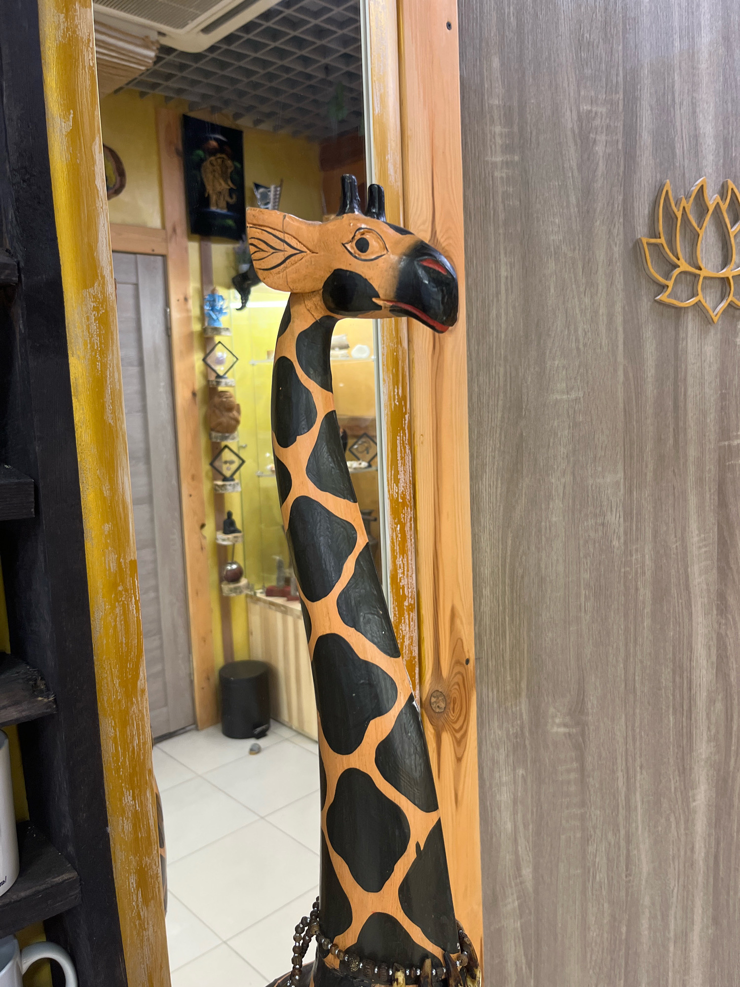The image shows a wooden giraffe statue with black spots and orange patches, positioned in front of a wooden door. To the left of the giraffe, a mirror reflects the interior of a room, which appears to be a shop or display area. The reflected space contains shelves with various decorative items, a black trash can, and a tiled floor. On the wooden door, a lotus flower motif is visible. The overall scene suggests an indoor environment, possibly a souvenir shop or a home decor store. The image does not provide any specific information about the city of Minsk, Belarus.