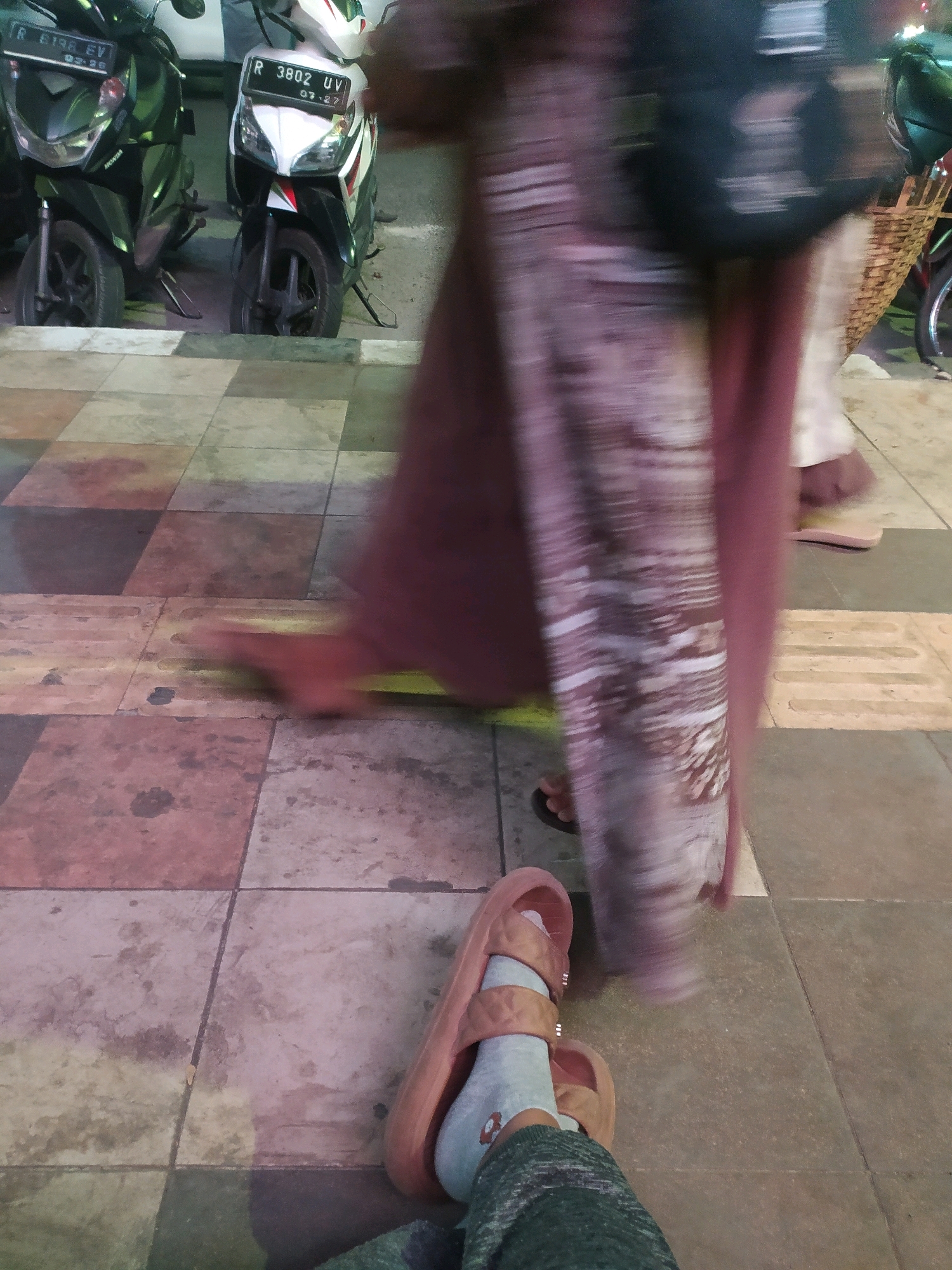 The image shows a street scene in Purbalingga, Indonesia, focusing on the ground and lower legs of several individuals. In the background, two scooters are parked, one black and one white, with license plates visible. One of the scooters has a red and white stripe detail on its front. The ground is paved with large square tiles in various shades of brown and gray, with some areas appearing wet or stained.
In the foreground, a person is wearing light grey socks and light brown slide sandals. The socks have a small red logo or design on them. The person's legs are clad in dark green, textured fabric, likely pants or leggings. The motion blur suggests movement within the frame. Another person's legs, wearing a long, flowing patterned garment (possibly a dress or long skirt) in muted colors, are also visible, with one foot in a light brown sandal. The pattern on the garment appears abstract or floral. A straw basket is also visible, slightly blurred, on the right side of the frame, possibly attached to a bicycle or another vehicle. The lighting suggests it is either dusk or nighttime.