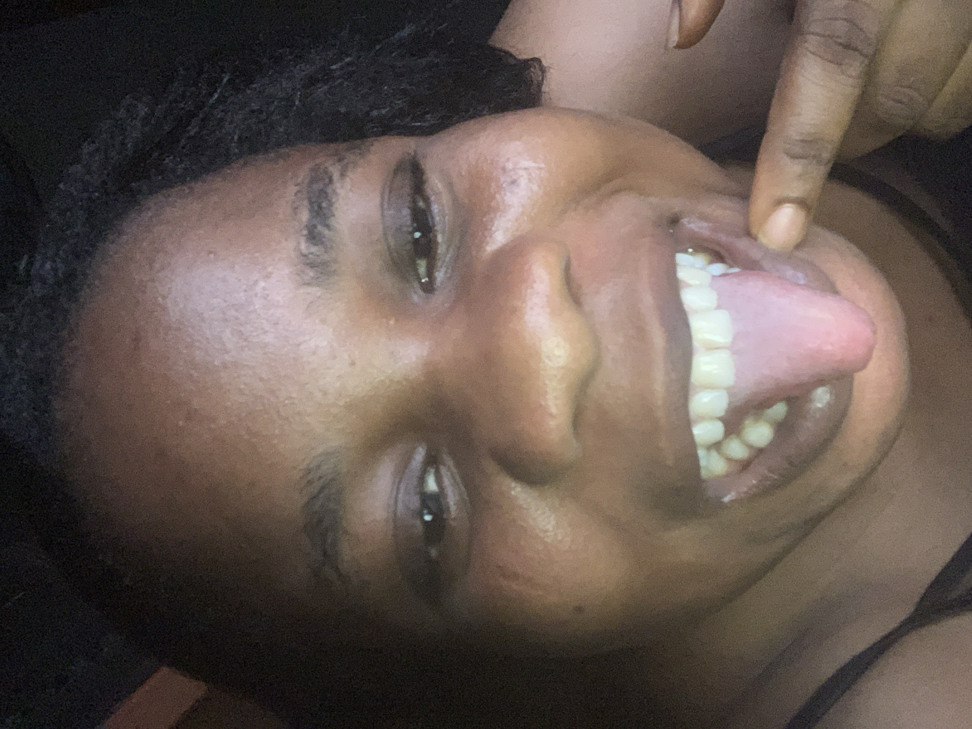 A close-up photograph shows a dark-skinned individual with curly black hair, lying on their back. Their eyes are open and focused, and their mouth is open with their tongue extended. One hand is visible, with a finger pointing toward the inside of their mouth. The background is dark and indistinct. The location is Unknown, Unknown.