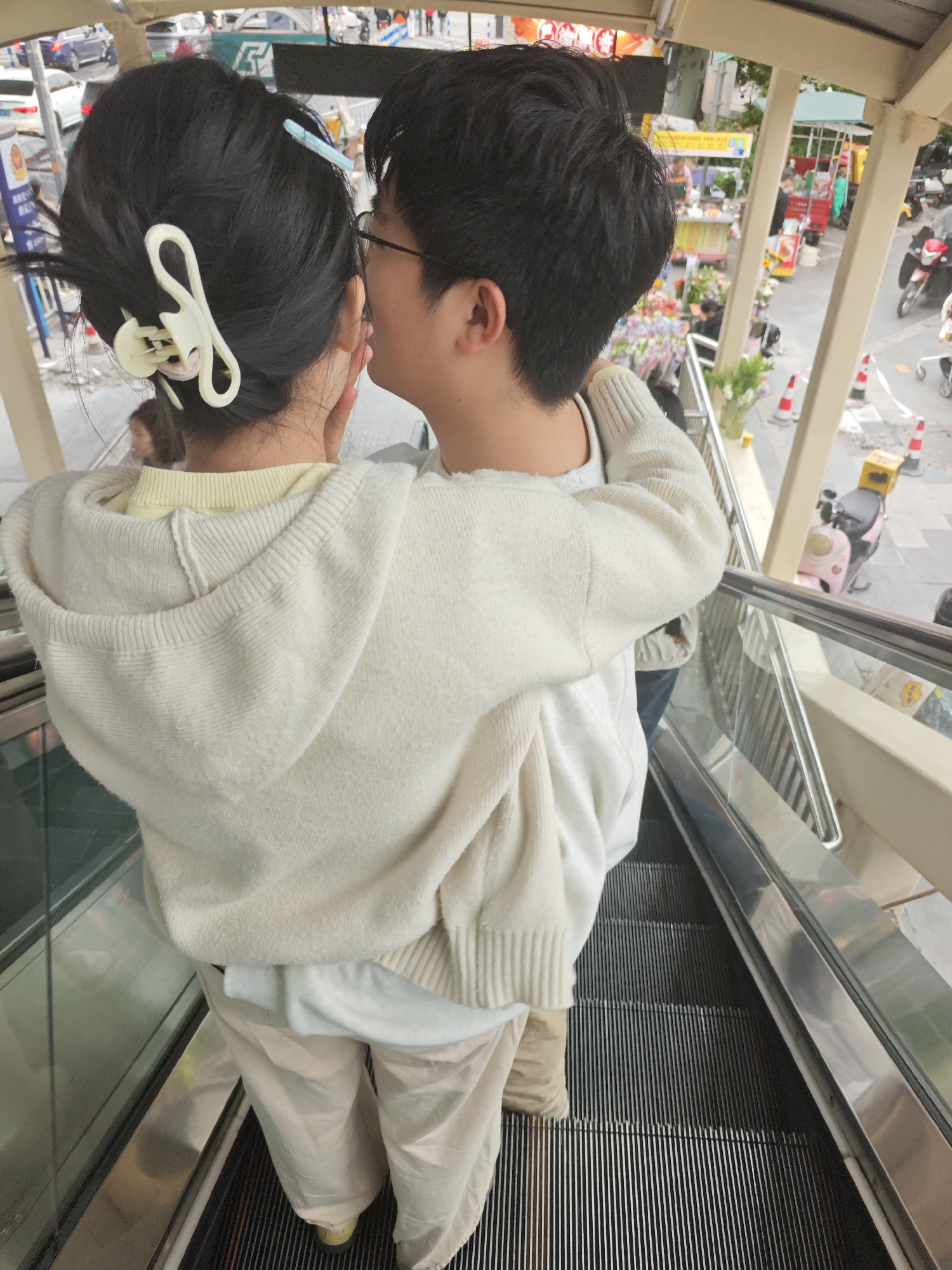 A young, dark-haired Asian couple, viewed from behind and above, stands on a descending escalator in the Chongqing High-tech Zone, China. The woman has a large white hair clip and a light blue clip holding her hair up, wears a light yellow shirt, and is wearing a large, off-white hooded sweatshirt. She has beige pants and is wearing gold-colored shoes. The man has dark hair, is wearing glasses, and is embracing the woman around her shoulders. The escalator is of standard commercial metal design, with a silver railing and a gray tread. Outside of the building there are traffic cones and a motor scooter.