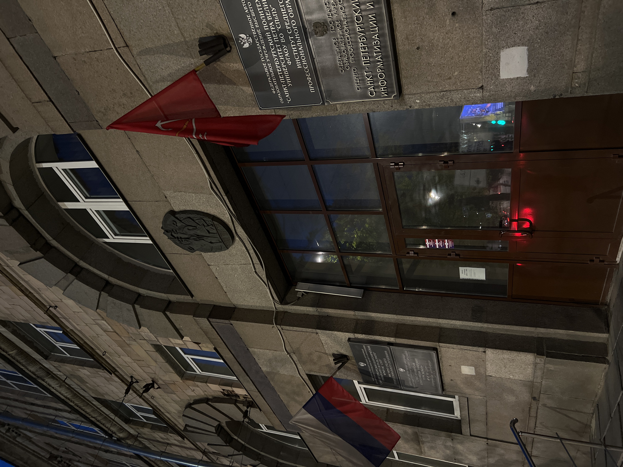The image shows the exterior of a stone building at night, with a red flag and a Russian flag displayed on the facade. The building has large windows and an arched entrance with a glass door. There are several signs on the wall, written in Russian, indicating that this is likely an educational institution. The overall atmosphere is somewhat austere and institutional.