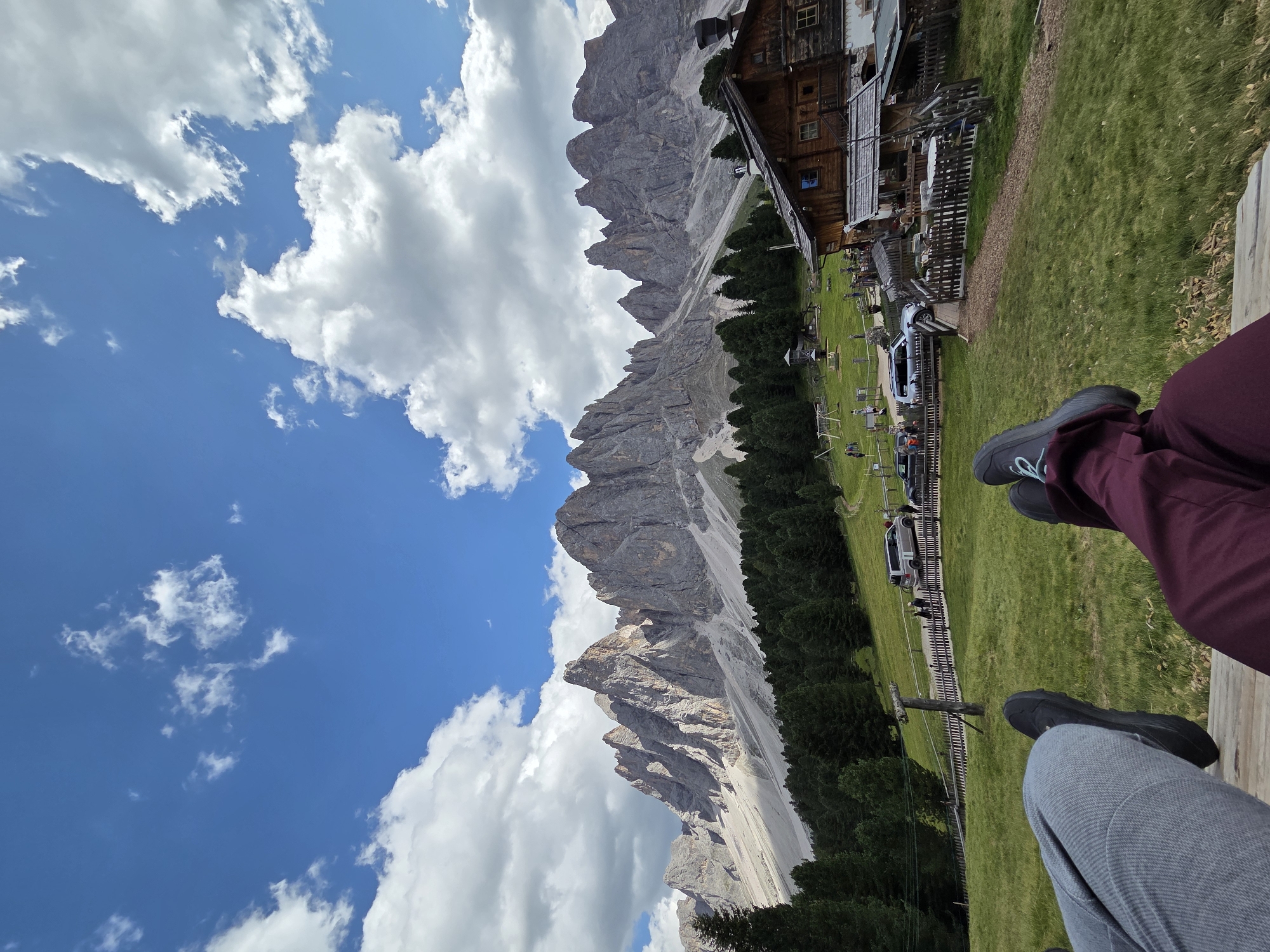 The image depicts a scenic mountain landscape with a rustic wooden building in the background. In the foreground, two people are seated on a wooden bench, their legs visible. One person is wearing burgundy pants and dark shoes, while the other is wearing grey pants and dark shoes.
The scene appears to be set in a mountainous region, possibly in Italy given the architectural style of the building which resembles a mountain hut or rifugio. The building has a traditional wooden facade with several windows. In front of the building, there is a grassy area with some wooden fences and tables, suggesting an outdoor seating area for the establishment. Several cars and people are visible in the middle ground, and a ski lift or similar transport system is also present, hinting at a location that might be active both in summer and winter.
The sky is a vibrant blue with scattered white, fluffy clouds, indicating fair weather. The sun is likely high in the sky, casting shadows that are not clearly discernible due to the angle of the image. The overall atmosphere seems relaxed and peaceful, with the individuals appearing to be enjoying the view. The rugged, grey mountain peaks dominate the background, showcasing a dramatic natural setting. The image is oriented vertically, with the sky occupying the left side and the mountain range extending diagonally from the bottom left to the top center.