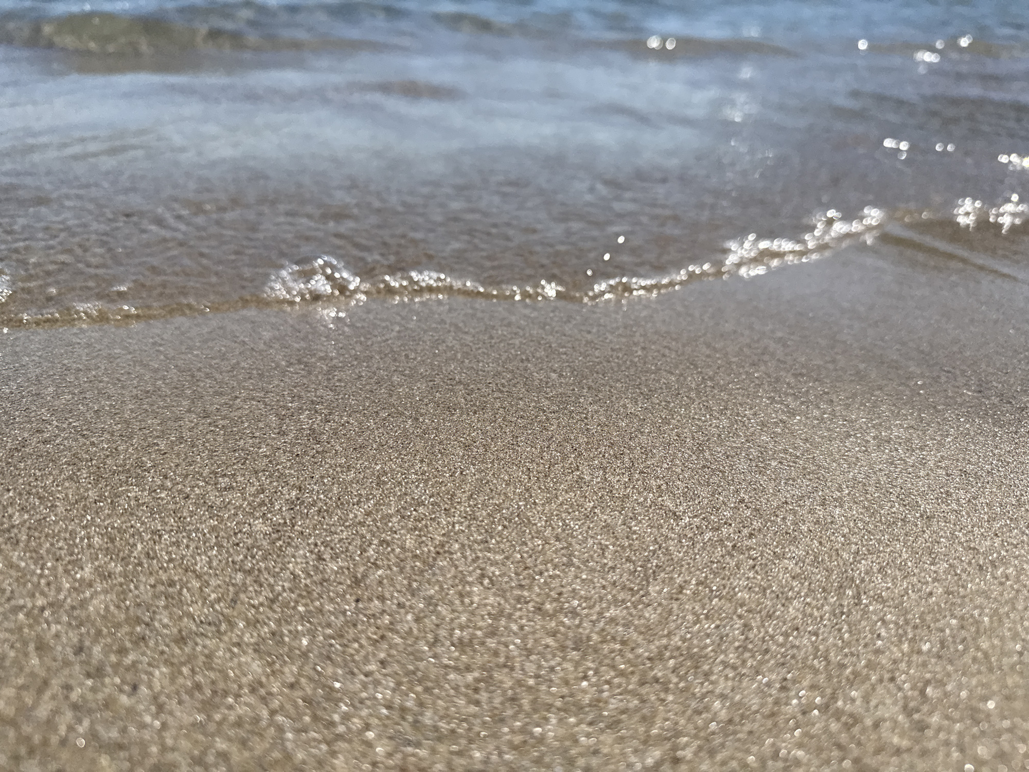 The image shows a close-up view of a sandy beach with the edge of the sea lapping at the shore. The sand is light brown and appears fine-grained. The water is a shallow wave, with small white foam and reflections of sunlight on its surface. The background shows more of the sea, with a blurry blue hue. The scene is likely located on one of the beaches in Sozopol, Bulgaria, such as the Central Beach or Harmanite Beach, known for their sandy shores and proximity to the town. There are no people or specific objects visible, only the natural elements of the beach and the sea.