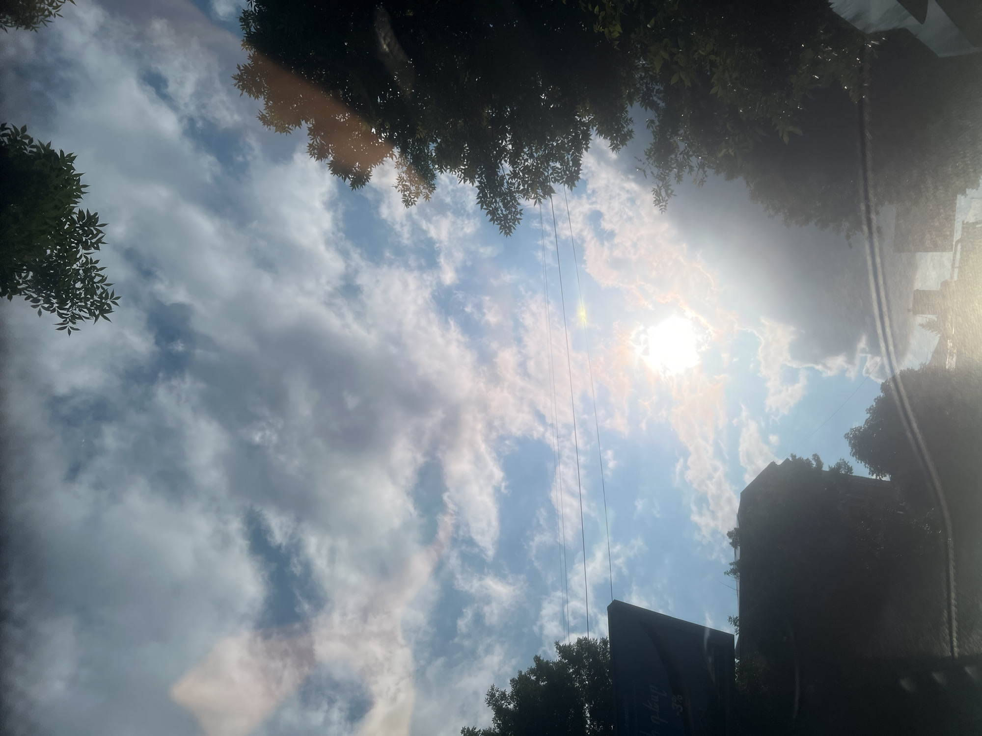 The image captures a view looking upwards, possibly from a window, showing a sky filled with scattered clouds. The sun is bright, casting a lens flare and illuminating the clouds, suggesting it is daytime. Silhouettes of trees frame the top and sides of the image, with some leaves visible. In the lower right quadrant, a dark, angular structure, possibly a building or sign, is visible in silhouette, with some faint text on its surface that is difficult to decipher. The scene appears to be an outdoor setting, with a mix of natural elements (sky, trees) and man-made structures. The overall mood is one of a bright, possibly warm day, with the sunlight creating a slightly hazy or glinting effect due to reflections. The orientation of the image is unusual, as if taken from a tilted perspective.