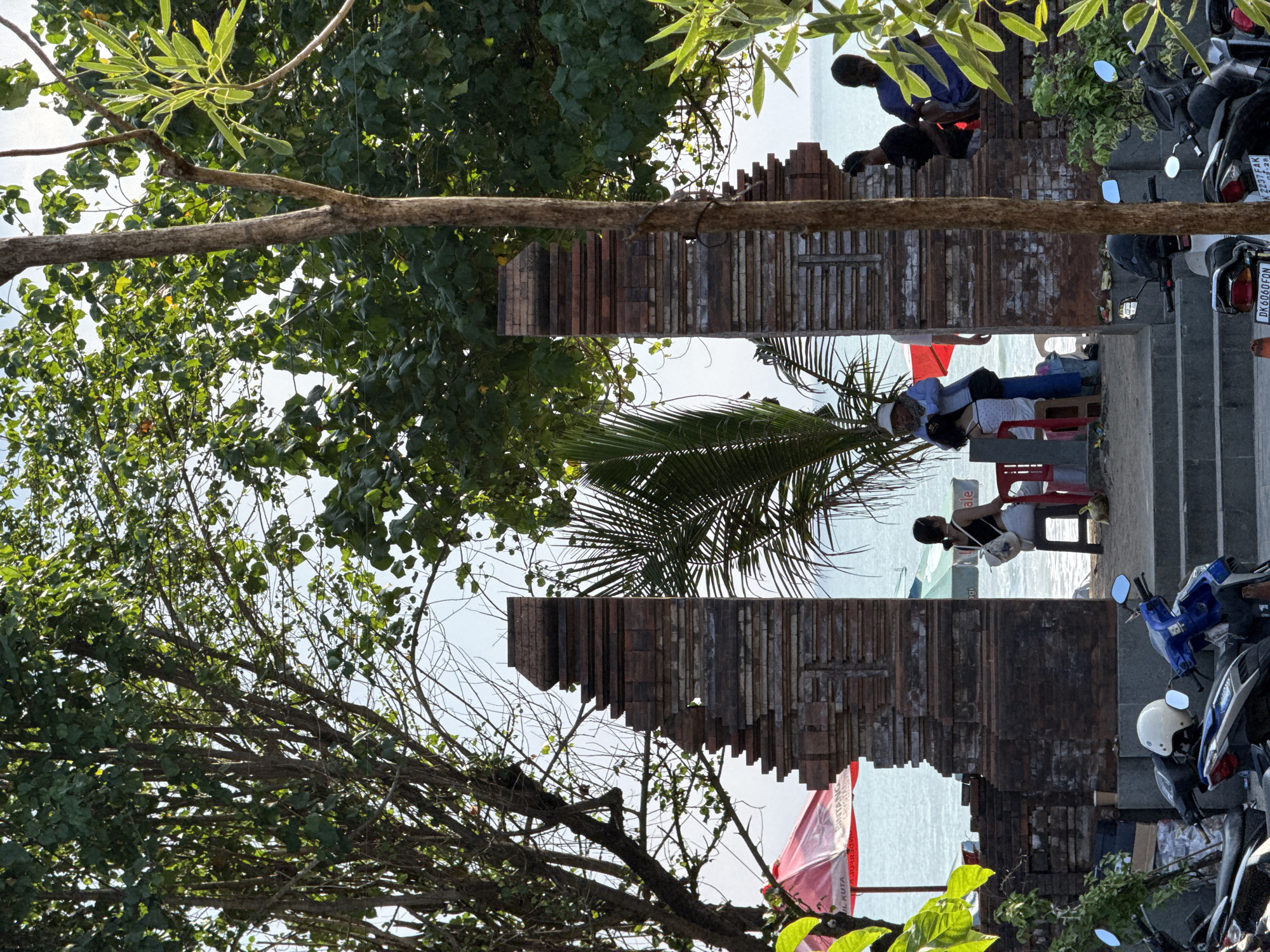 This media file captures a vibrant outdoor scene, likely a coastal or beachside area in Kuta, Indonesia. The image is dominated by lush green foliage from several trees that frame the view, with a thick branch extending horizontally across the mid-ground.
In the center, two distinct, reddish-brown and greyish-brown brick or tile-like structures, resembling decorative walls or gateposts with uneven tops, create a visual corridor leading towards a bright expanse of ocean or sea in the background.
Several people are visible within the scene, engaged in leisure activities. On the right, two individuals, one in a blue shirt and another in a black shirt, are seated on steps or a low wall near parked motorcycles. Further to the left and closer to the water, a person wearing a white cap, light blue top, and white shorts is seated on a red plastic chair, appearing to relax and enjoy the view. Another person is partially visible near the water, also on a red chair. To the far left, a red umbrella is visible near the water's edge.
The scene suggests a bright, sunny day, with clear skies illuminating the landscape. Several motorcycles are parked on the right, some with white helmets. Visible text includes a motorcycle license plate reading "DK 6060 FON," and another partially visible plate "K 2275 AK," both typical of Bali, Indonesia. A red umbrella displays the text "BINTANG CRYSTAL" and "KUTA," further confirming the location and suggesting a beachside establishment. The overall atmosphere appears relaxed and inviting, characteristic of a popular tourist destination.