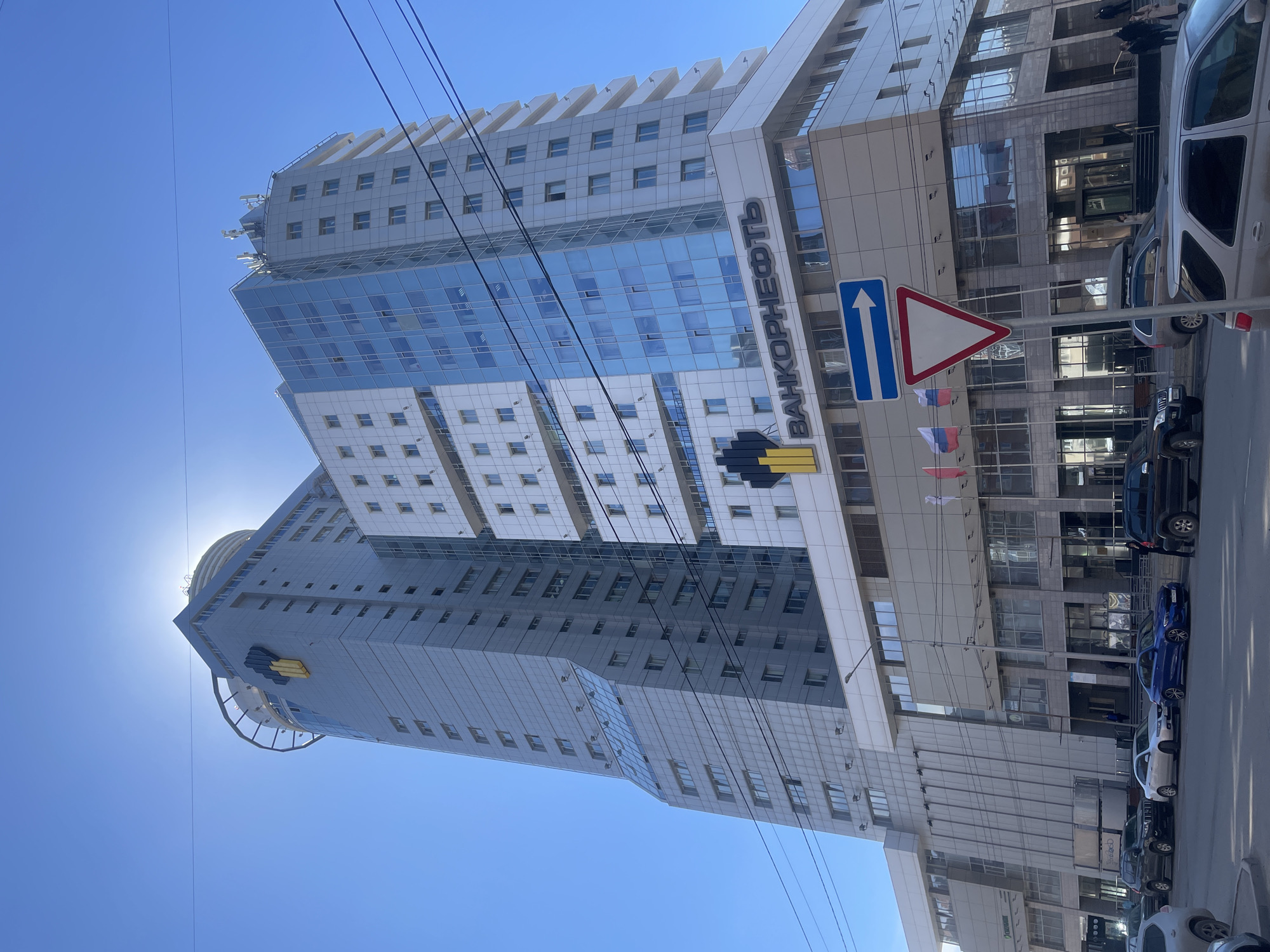 This bright, sunny daytime image captures a street view dominated by two modern high-rise buildings in Krasnoyarsk, Russia. The building on the right features a facade of light-colored panels and reflective blue-tinted glass windows. A prominent vertical sign on this building displays "БАНКОРНЕФТЬ" (BANKORNEFT) in Cyrillic lettering, accompanied by a distinct yellow and black corporate logo, possibly resembling an abstract representation of an oil derrick or flame. Several small Russian flags are visible hanging near the building's entrance at street level.
The second building, situated slightly behind and to the left, is also a tall, contemporary structure with a similar gray and glass aesthetic. The bright sun is positioned directly behind this building's upper section, creating a strong sunburst effect against the clear blue sky. A distinctive circular or cylindrical architectural feature is visible at the very top of this building.
At street level, a line of cars is parked along the curb in front of the buildings. A few pedestrians can be seen near one of the entrances of the rightmost building, suggesting everyday activity. Two road signs, a blue rectangular "straight ahead" arrow and a red triangular "yield" sign, are prominently displayed near the street. Numerous dark power lines crisscross the upper portion of the clear blue sky. The scene depicts a typical urban environment on a clear, sunny day.