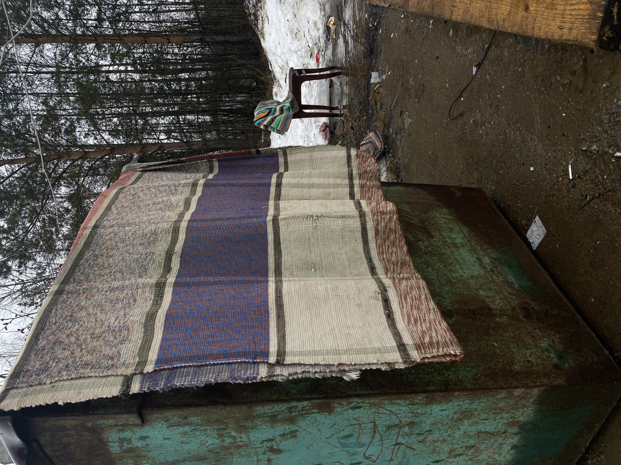 The image depicts a scene in what appears to be a forest clearing in Ol'khovskoye, Russia. In the foreground, a large, weathered metal dumpster is partially covered by a striped woven rug or blanket. Behind the dumpster, in the mid-ground, a wooden chair sits near a patch of melting snow. A colorful blanket or garment is draped over the chair's back. The ground is muddy and littered with debris. In the background, a dense stand of trees stretches upwards, their branches bare against an overcast sky. The overall impression is one of neglect or abandonment.
