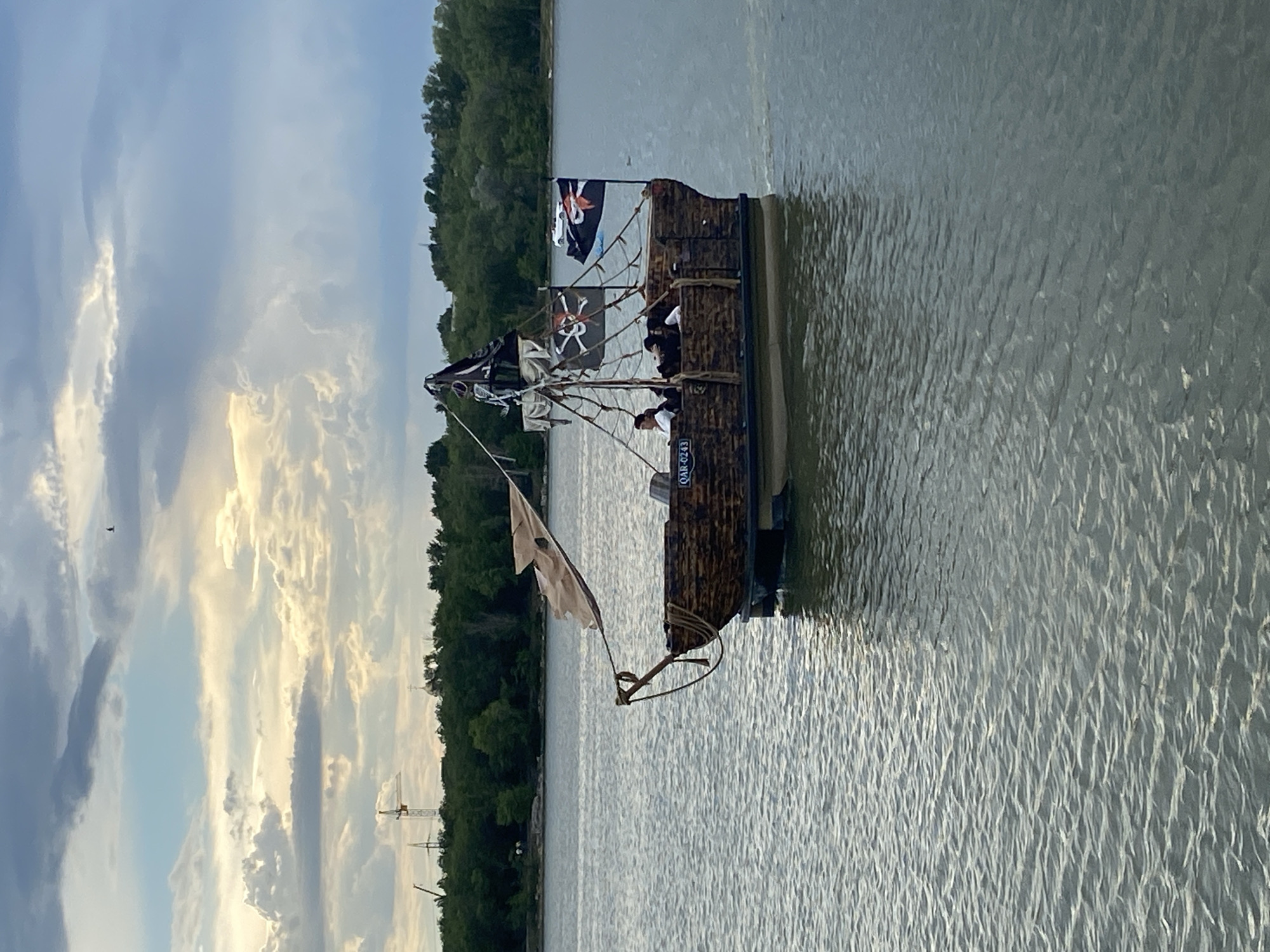 A low-angle, full shot captures a replica pirate ship with a wooden hull on a body of water. The ship has two masts with tattered sails and is adorned with two skull and crossbones flags and another black flag. Several people are visible on deck, some dressed in white shirts and hats, suggesting a themed event. The ship appears to be moving, with a slight wake visible behind it. The background features a dense line of green trees, a crane, and a church steeple, possibly indicating a city or town adjacent to the water. The sky is partly cloudy with patches of sunlight breaking through, creating a dramatic atmosphere. The water is choppy, reflecting the sky and surrounding elements. A small registration number "QAR-0243" is visible on the side of the boat. The overall scene evokes a sense of adventure and historical reenactment.