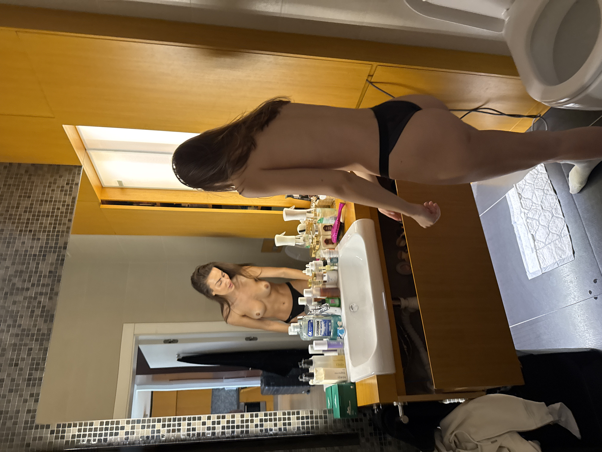 A full shot captures a woman standing in a bathroom, her back to the camera, with her reflection visible in a large mirror. She is wearing dark underwear and appears to be topless. Her long, dark hair cascades down her back.
The bathroom features light brown wooden cabinets above and below a large mirror. A white sink with various bottles of toiletries, including Listerine, is visible in the reflection. To the right of the woman, a white toilet is partially visible. The floor is covered with dark tiles, and a white, textured mat is placed in front of the toilet. On the far left, a tiled wall with small, square grey and white tiles can be seen. The overall lighting suggests an indoor, possibly evening, setting. The room appears tidy and well-maintained.