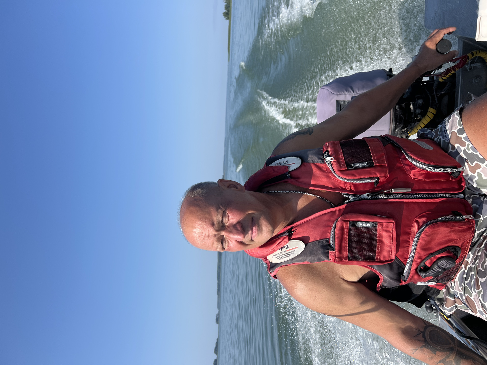 A bald, middle-aged Caucasian man wearing a red life vest and camouflage shorts is the primary subject. He is on a boat on a body of water, with a wake trailing behind. The man is looking to his left, and appears to be holding the boat's tiller with his right hand. The background consists of a clear blue sky and a distant shoreline with vegetation. The image appears to be taken during the daytime, under clear weather conditions, and it is likely the man is in Beregovoe, Russia.