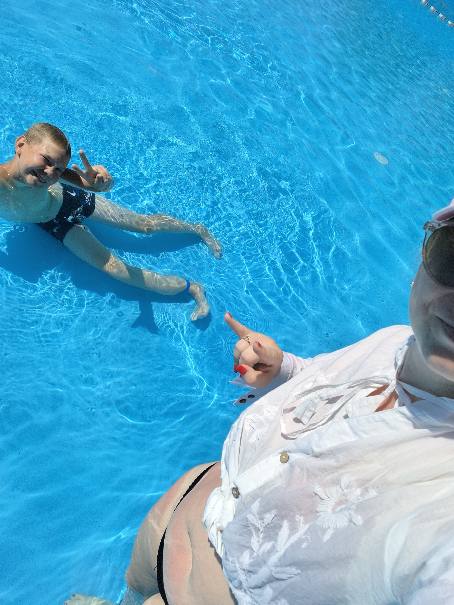 In a swimming pool in Anapa, Russia, a young boy in black swimming trunks floats on his back, his legs extended and submerged in the clear blue water, and he smiles while holding up two fingers in a peace sign. To the right, a person wearing a white, open, embroidered shirt and sunglasses, with red nail polish, extends their right hand and points forward. The water is bright and reflects sunlight, creating ripples and shadows.