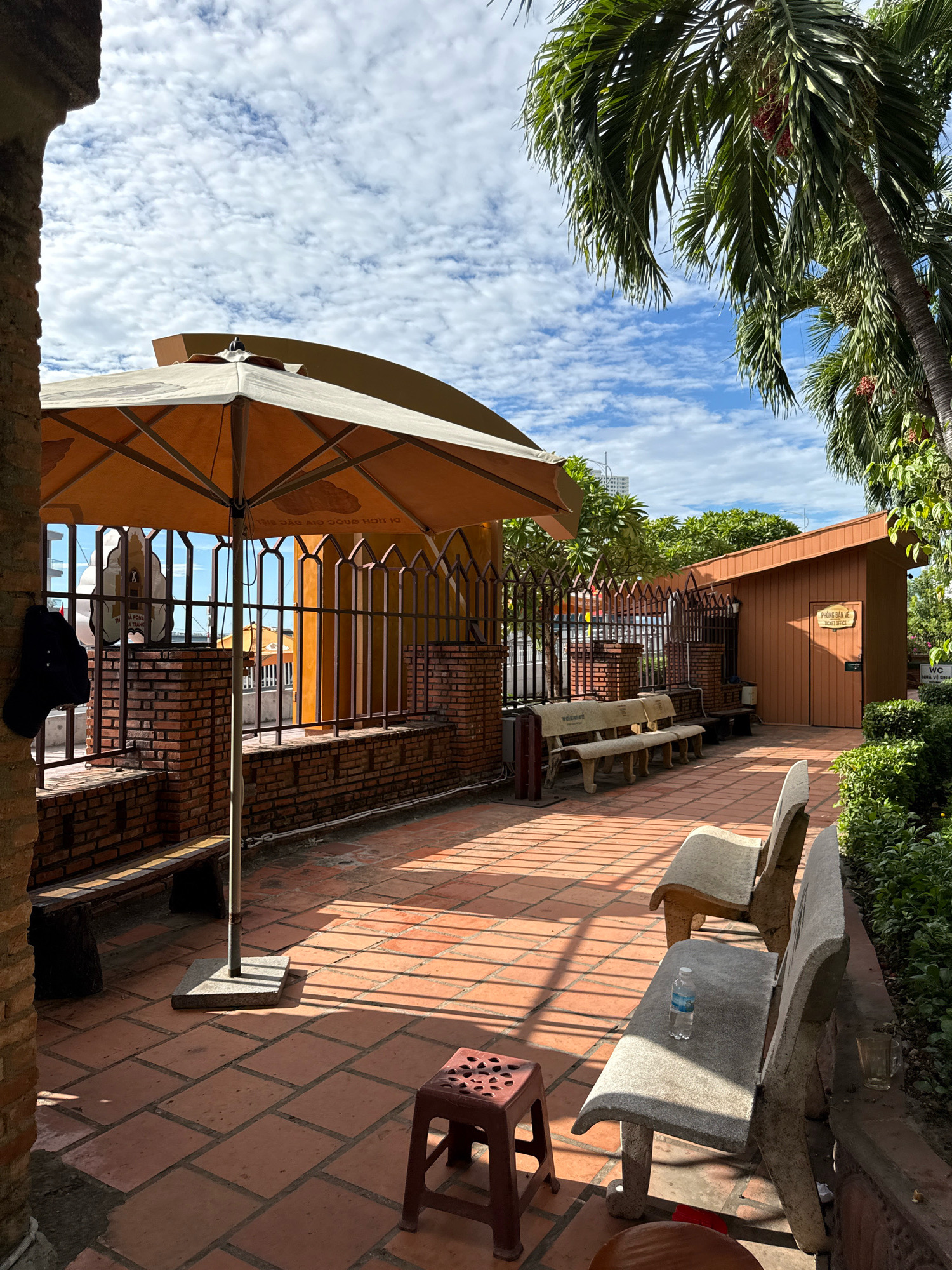 The image depicts an outdoor seating area with terracotta tiled flooring, characteristic of many establishments in Nha Trang, Vietnam. The scene is bathed in sunlight, casting distinct shadows from an overhead umbrella and a wrought-iron fence. Several concrete benches are arranged around the space, some with visible water bottles and a glass, suggesting recent use. In the background, a small wooden building with a sign reading "PHÒNG BÁN VÉ - TICKET OFFICE" indicates a ticket sales point, commonly found at tourist attractions or transportation hubs in cities like Nha Trang. Palm trees frame the upper right portion of the image, reinforcing the tropical setting. The sky is a clear blue dotted with white clouds. No individuals are present in the frame.