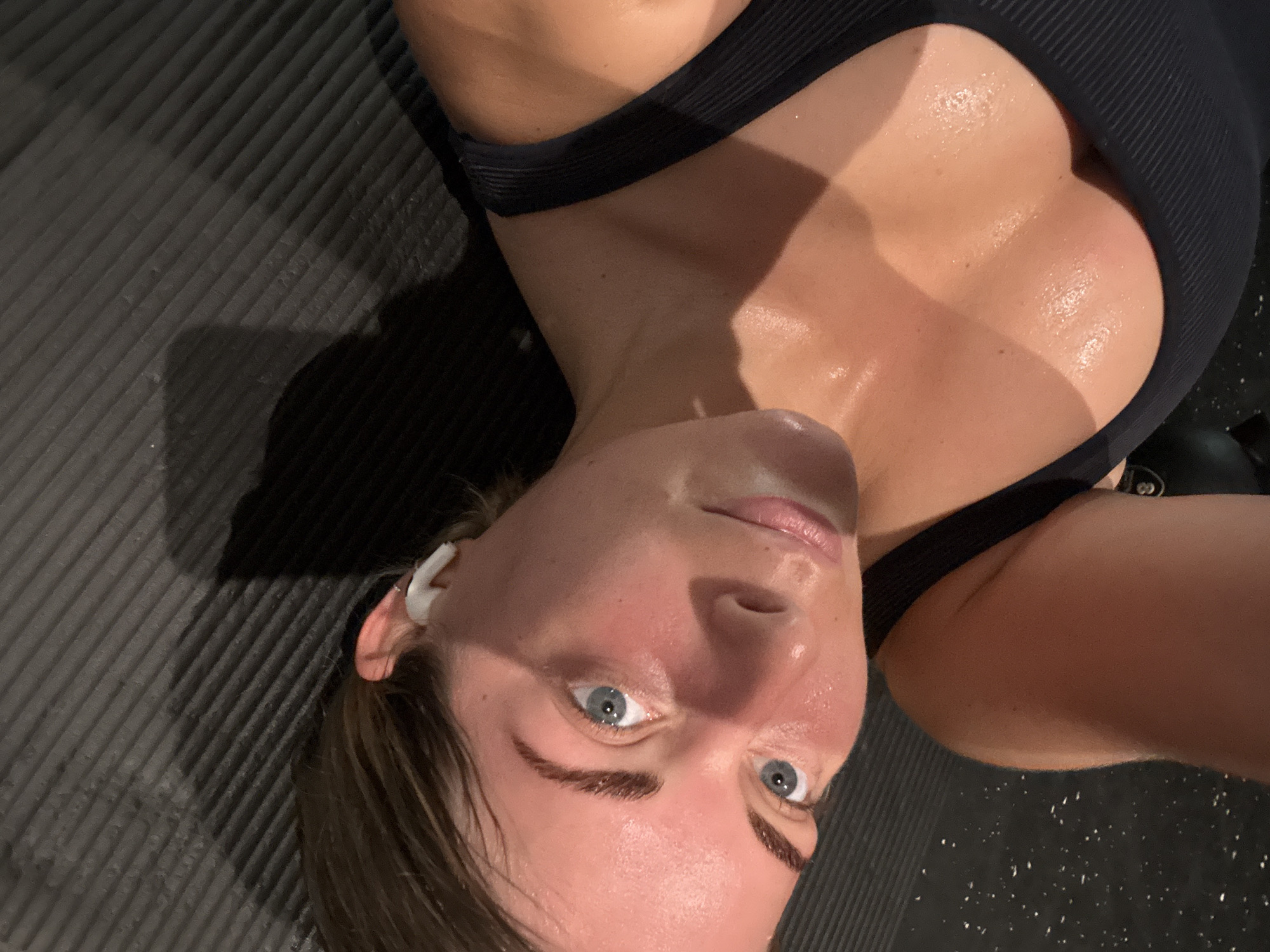 A close-up, upside-down selfie captures a woman with fair skin and blue eyes, presumably after a workout. Her face is flushed, and beads of sweat glisten on her forehead, nose, and chest. She wears a black sports bra with a ribbed texture, offering a partial view of her upper chest and shoulder. A white wireless earbud is visible in her left ear. The background consists of a dark grey textured surface, potentially a gym mat or flooring. There is no evidence in the image to suggest that the location is Paris, France.
