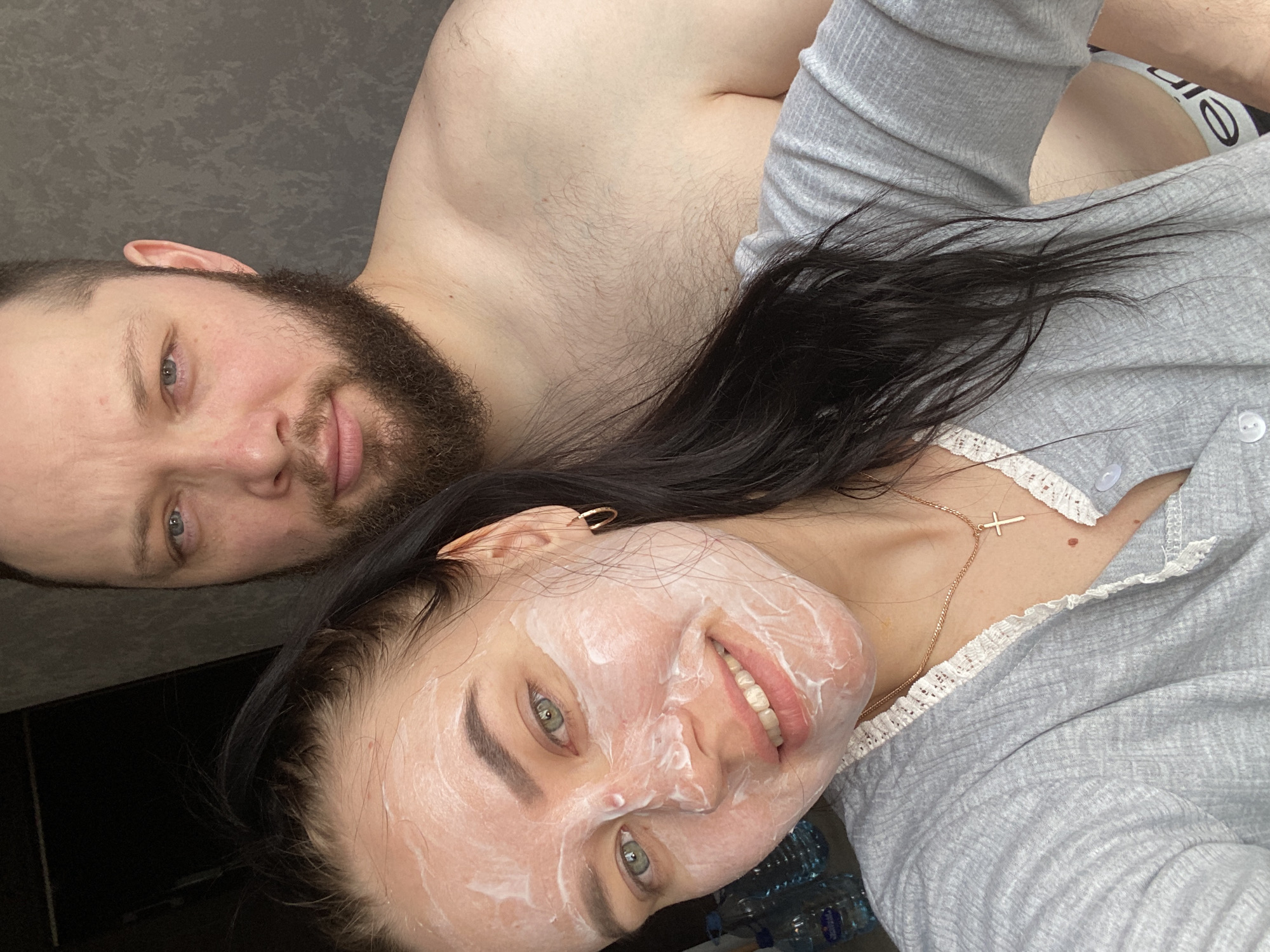 A selfie captures a man and a woman. The man, positioned in the upper left, has a beard and is shirtless. The woman, in the foreground, has a white facial mask applied and wears a gray shirt with a lace collar and a gold cross necklace. Dark hair frames her face. In the lower right, partially visible are water bottles. An underwear brand label with text is visible in the top right of the image.
The location is in the city of Chaikovsky, Russia.