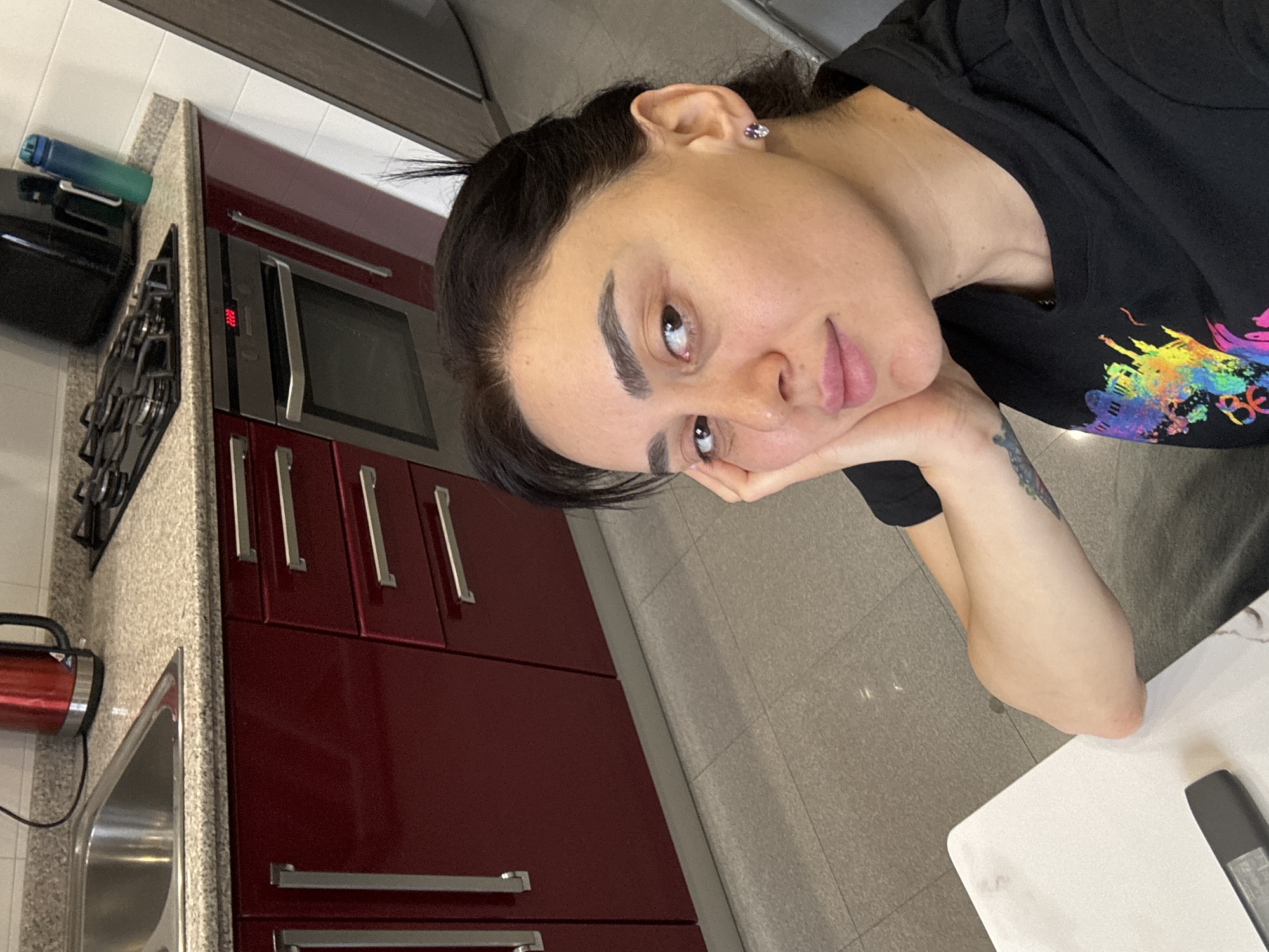 A woman is posing for a selfie in a kitchen. She is wearing a black t-shirt with a colorful design. Her hair is dark and pulled back. She has an earring in her left ear and a tattoo on her left forearm. The kitchen features red cabinets with silver handles and a gray countertop. A sink, stovetop, and oven are visible. The location appears to be in Tbilisi, Georgia based on the architecture and interior design.